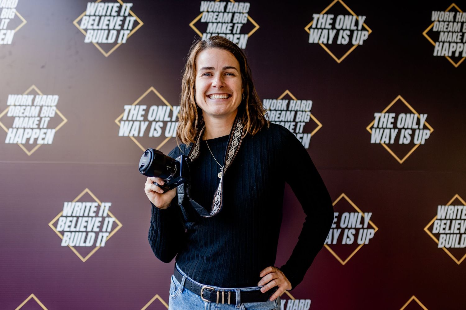 A smiling photographer holding a camera in front of a motivational quote backdrop at an event.