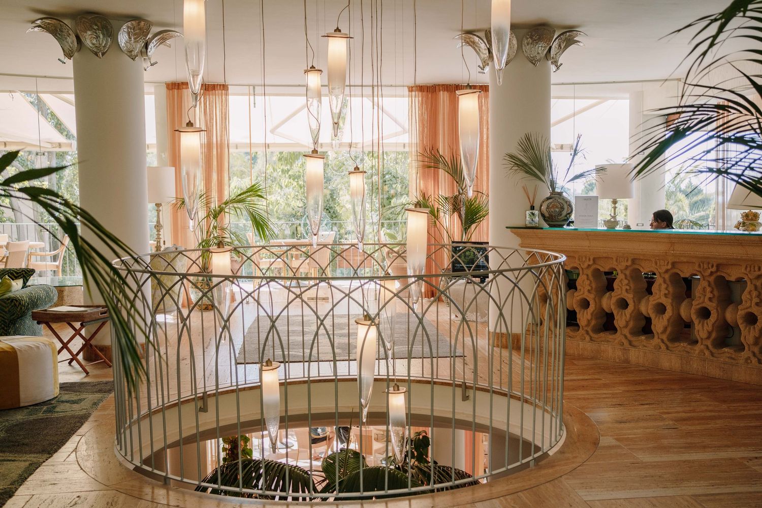 Elegant hotel lobby with a curved glass balustrade, hanging pendant lights, tropical plants, and a wooden reception desk on polished floors.