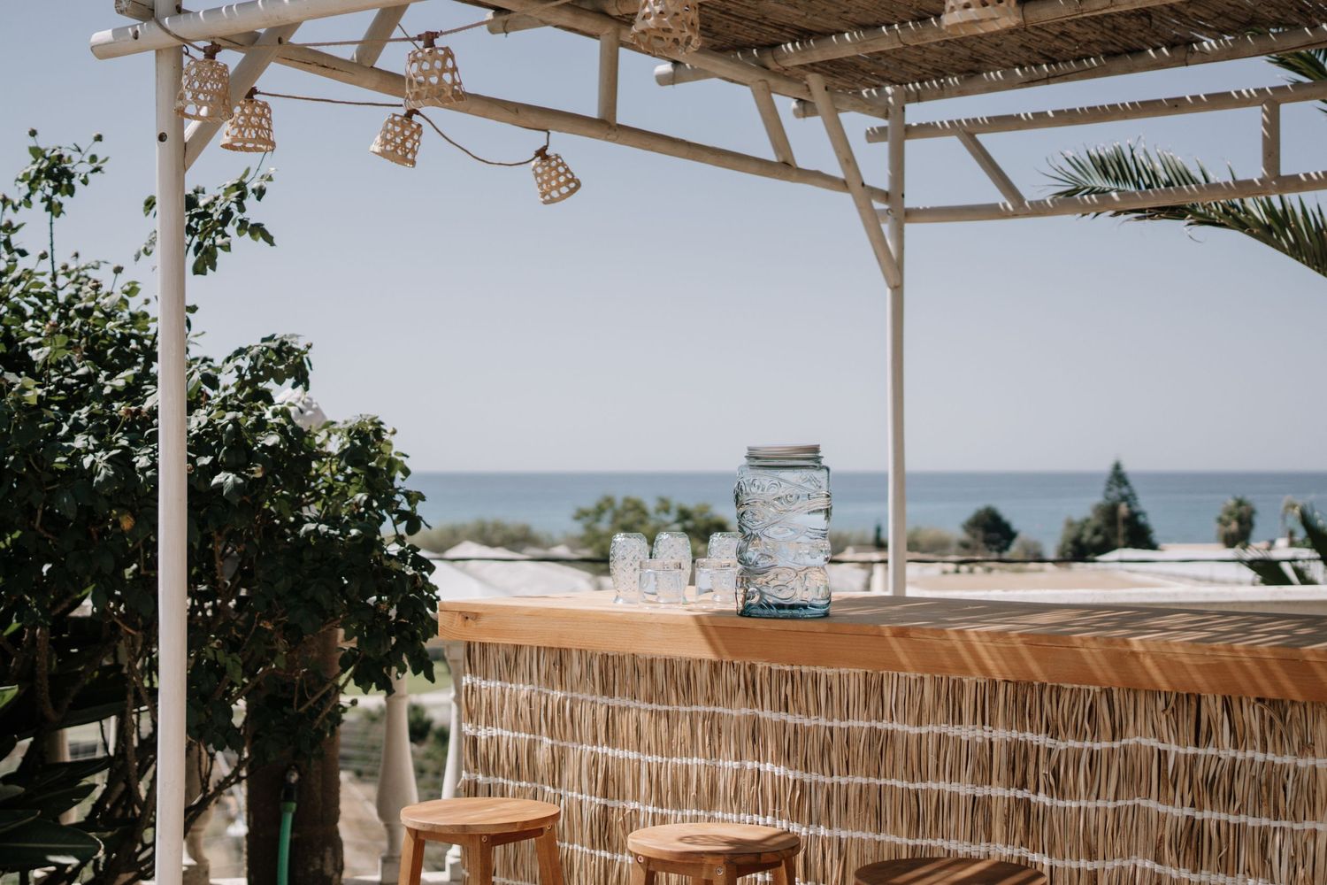Beachside tiki bar with rattan stools, mason jar decor, and ocean views under a white pergola.