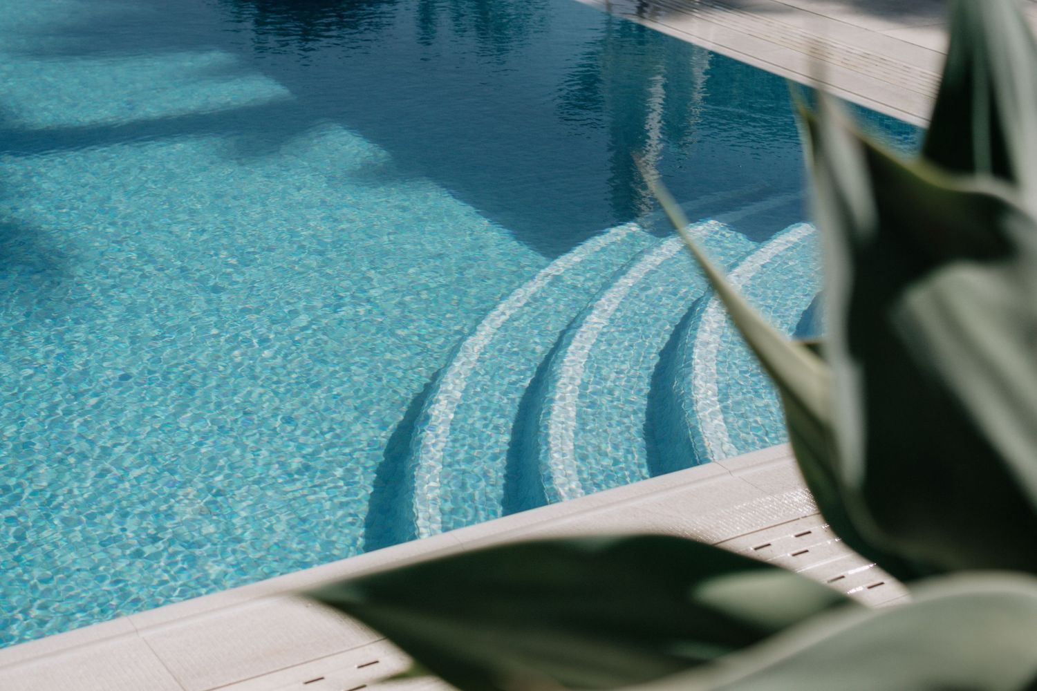 A serene luxury swimming pool with clear blue water, tiled steps, and lush green tropical leaves in the foreground.