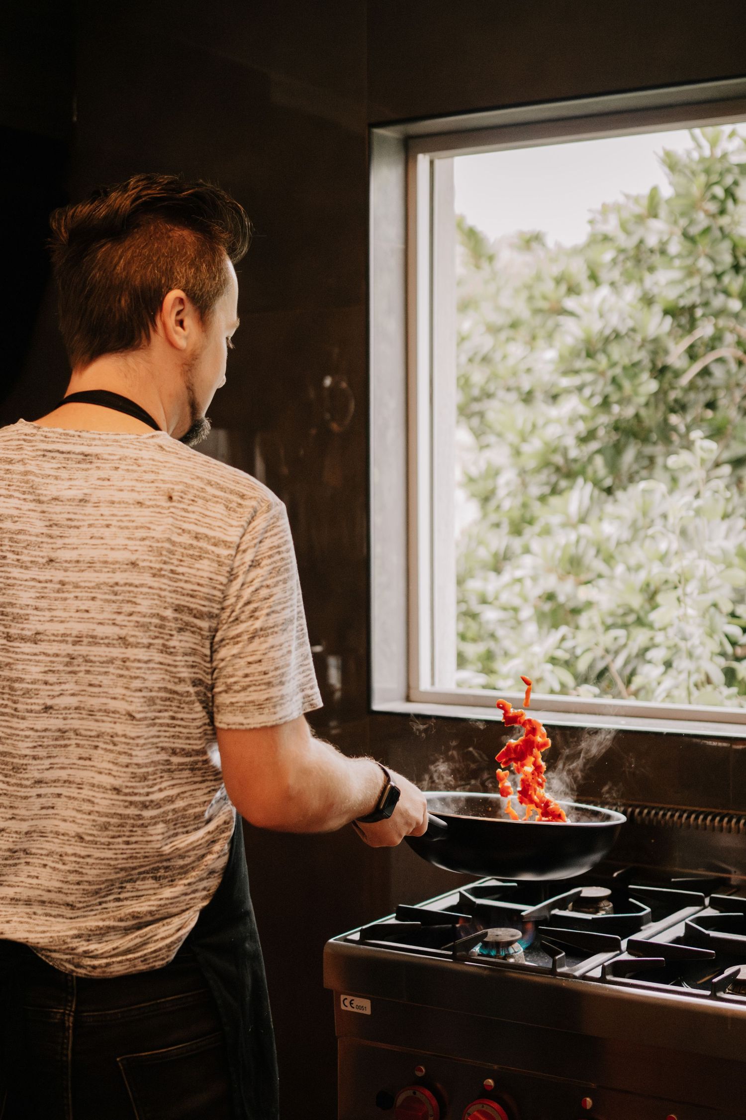 Man sautéing red peppers in a pan on a gas stove near a window with green trees outside.