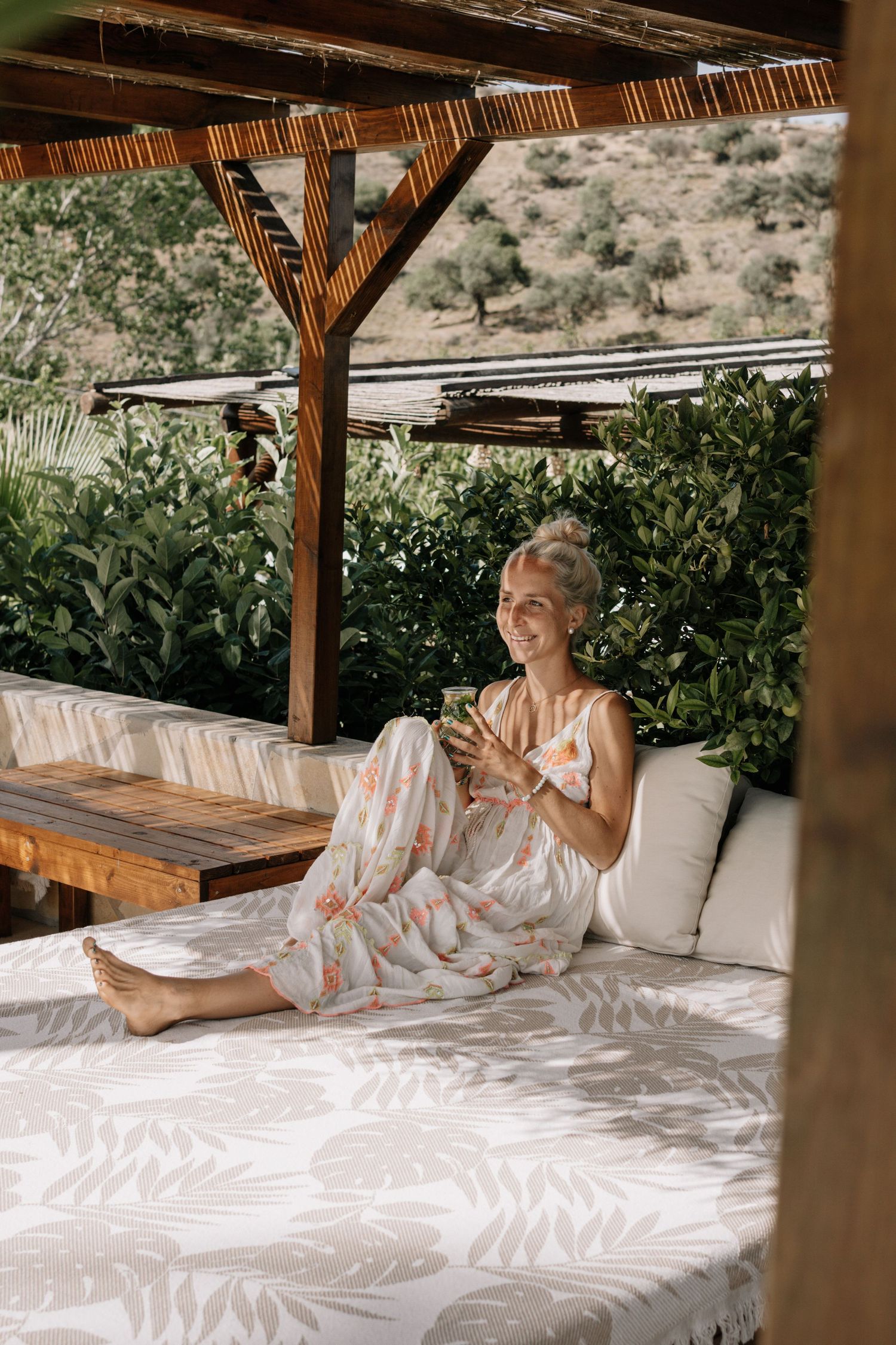 A smiling blonde woman in floral pajamas relaxes on an outdoor daybed, holding a drink surrounded by lush greenery.