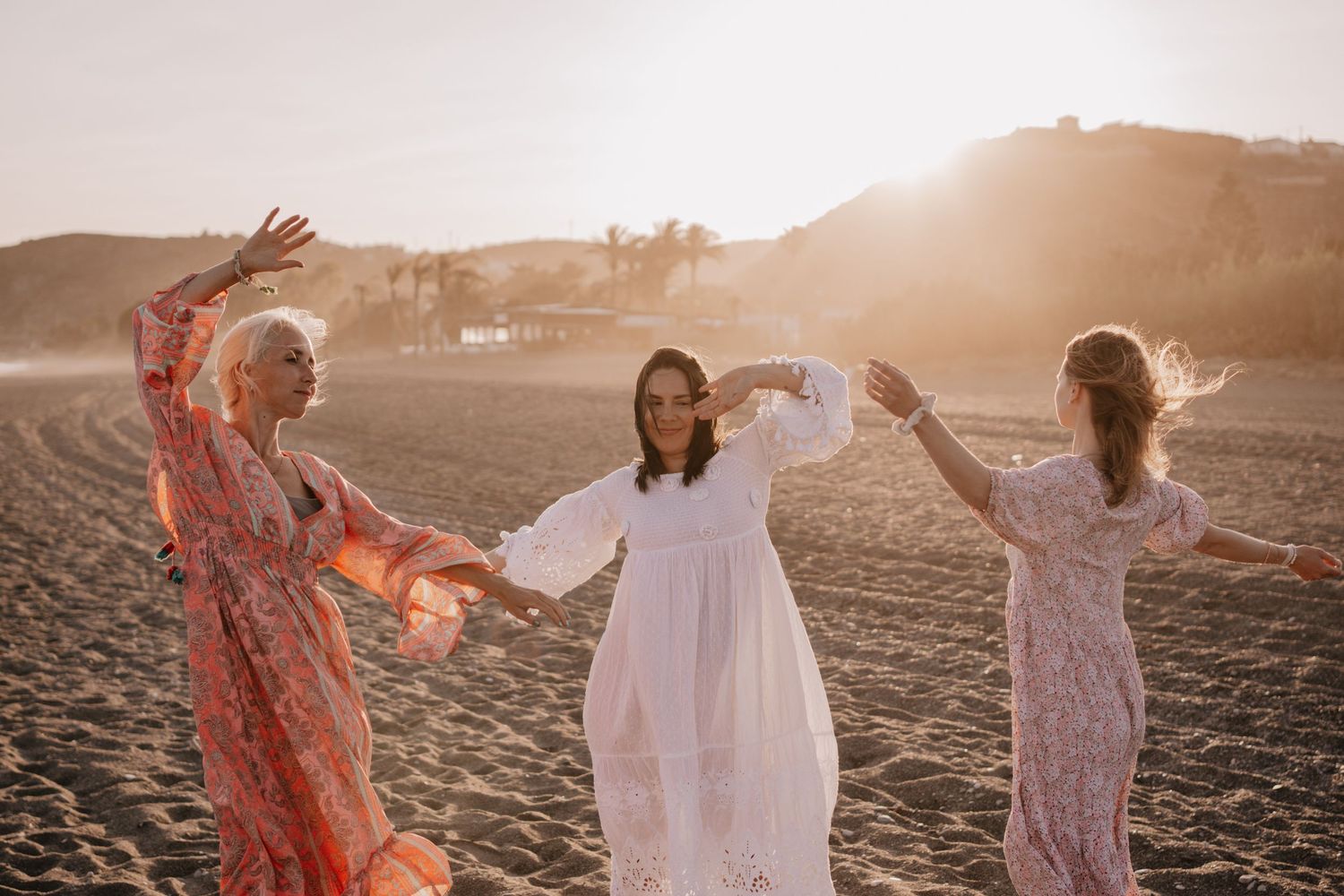Three women in flowing dresses dance joyfully on a beach at golden sunset.