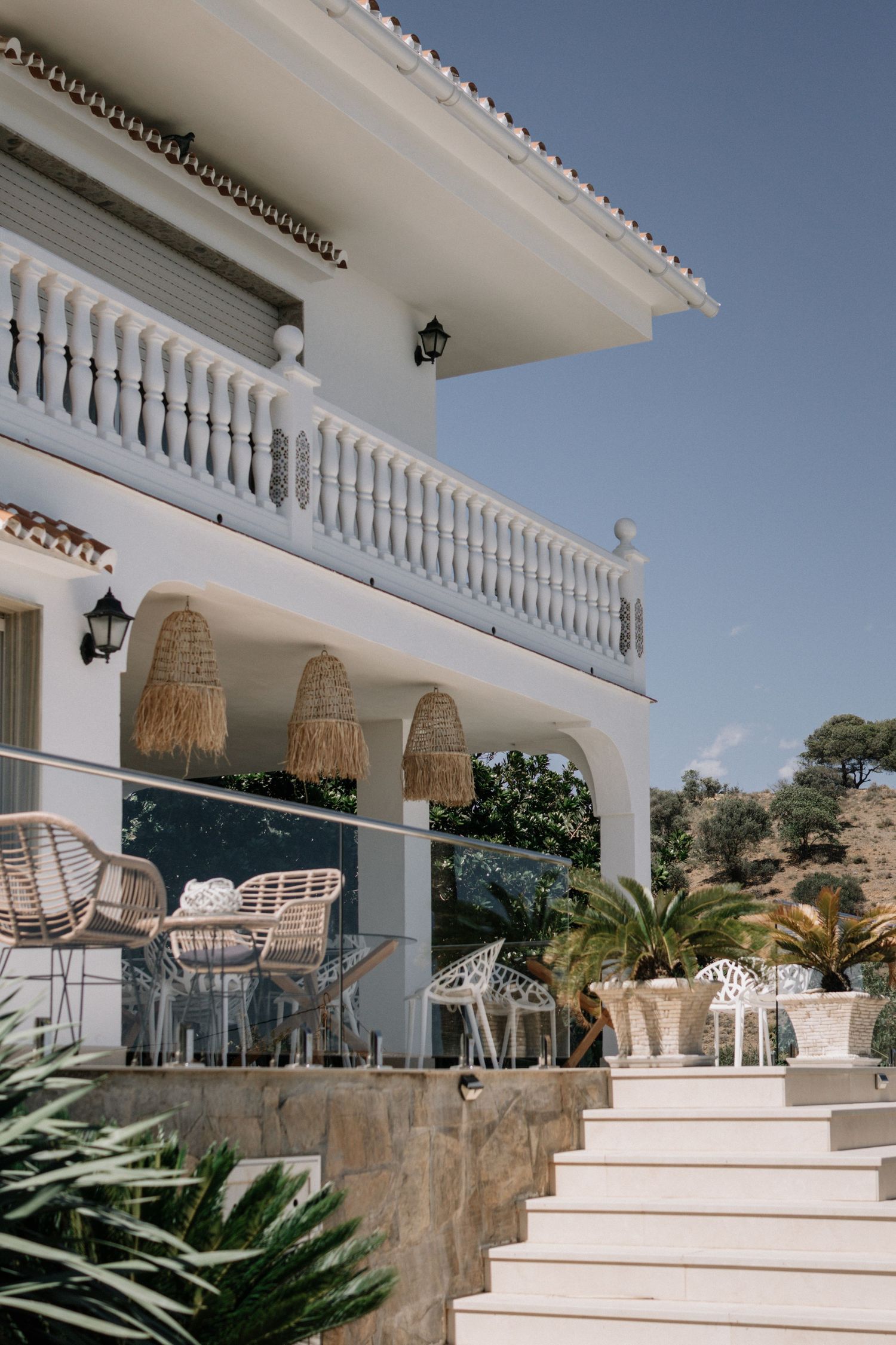 White Mediterranean villa with rattan chairs, stone steps, balcony, and palm trees under a clear blue sky.