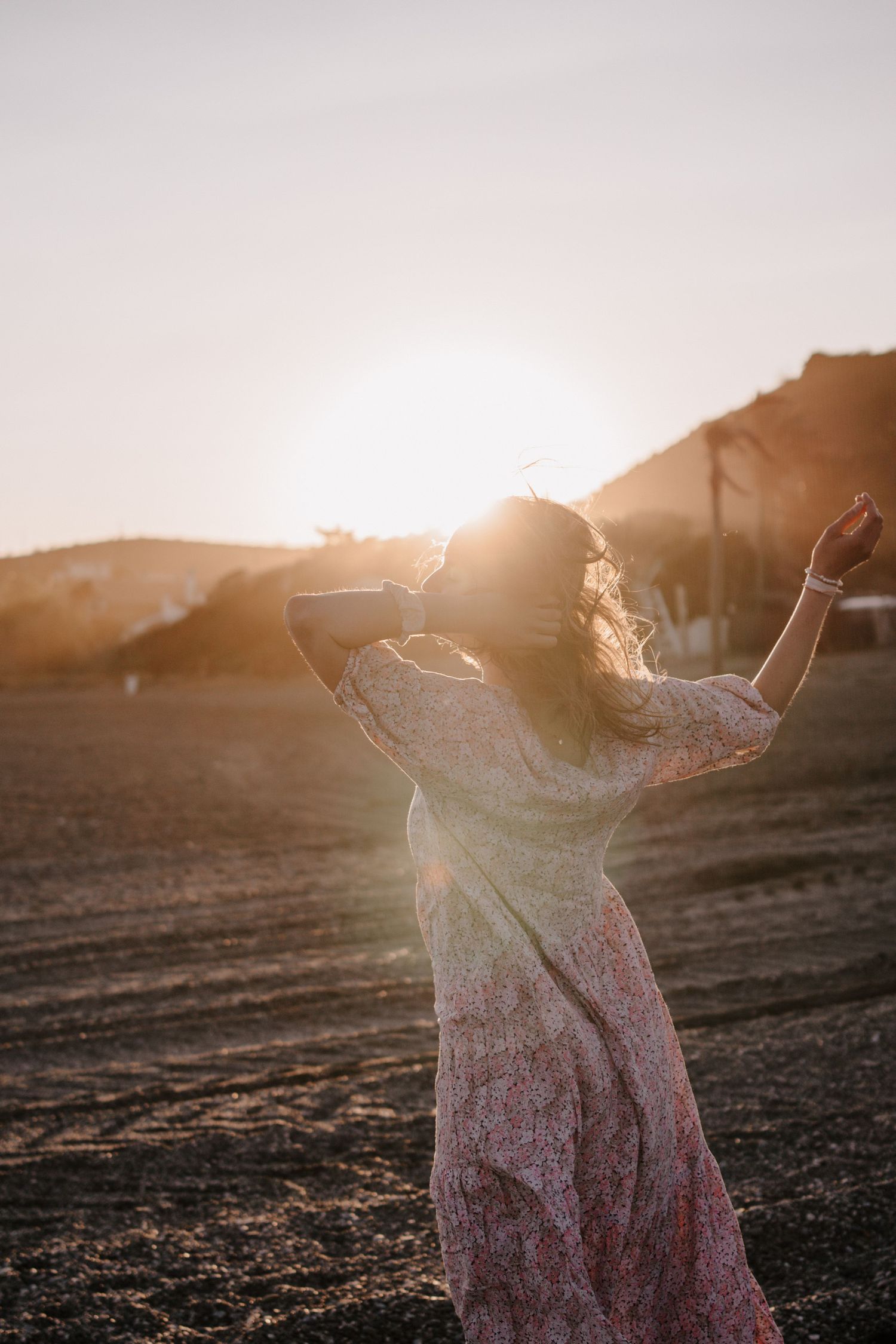Woman in floral dress dancing at sunset in an open field with golden backlight and mountain silhouette.