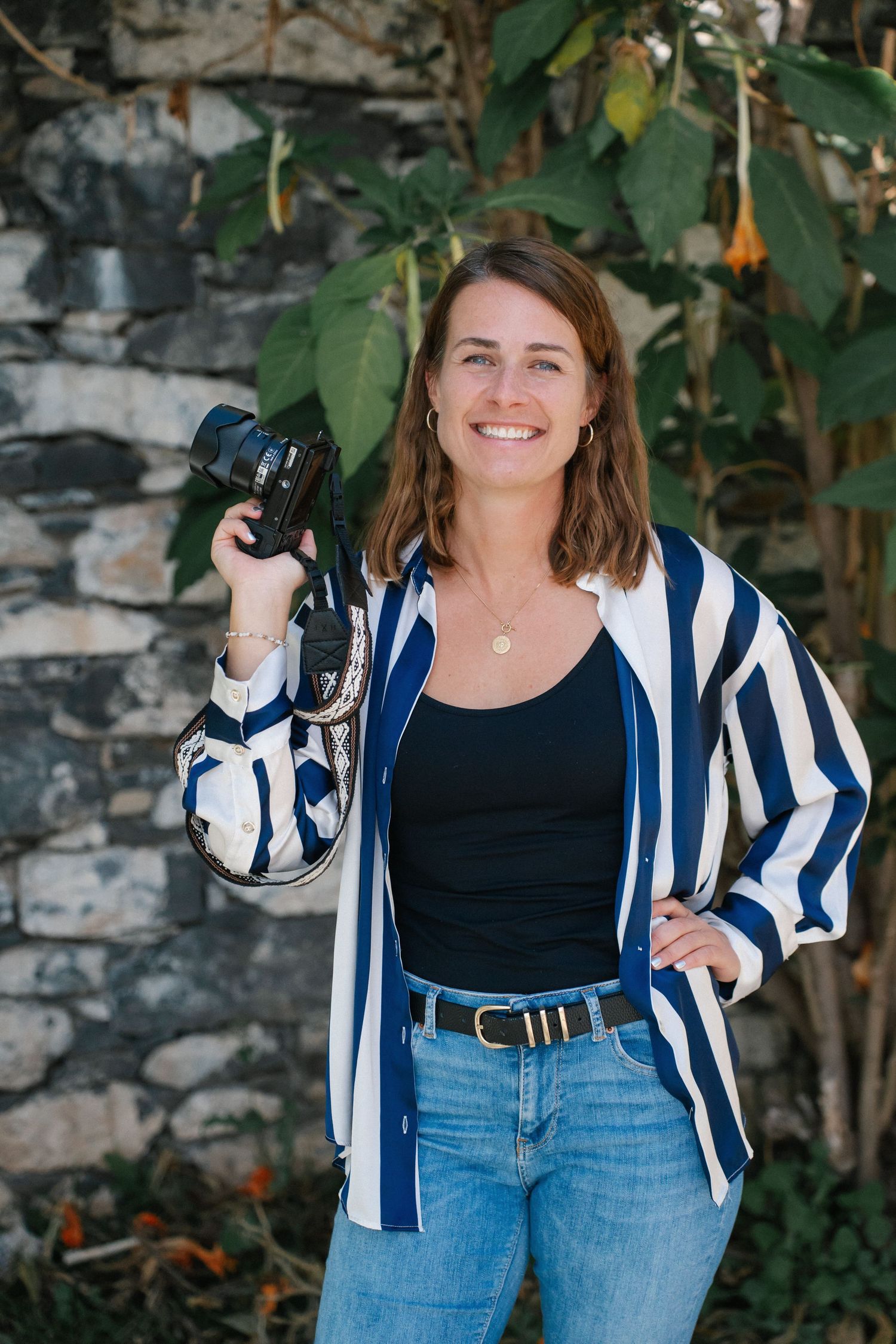 Smiling brunette woman holding camera outdoors against stone wall with green foliage.