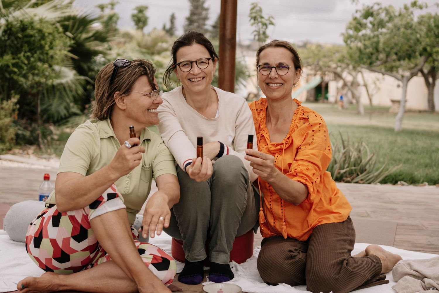 Three smiling women sitting outdoors on blanket holding cosmetic products.