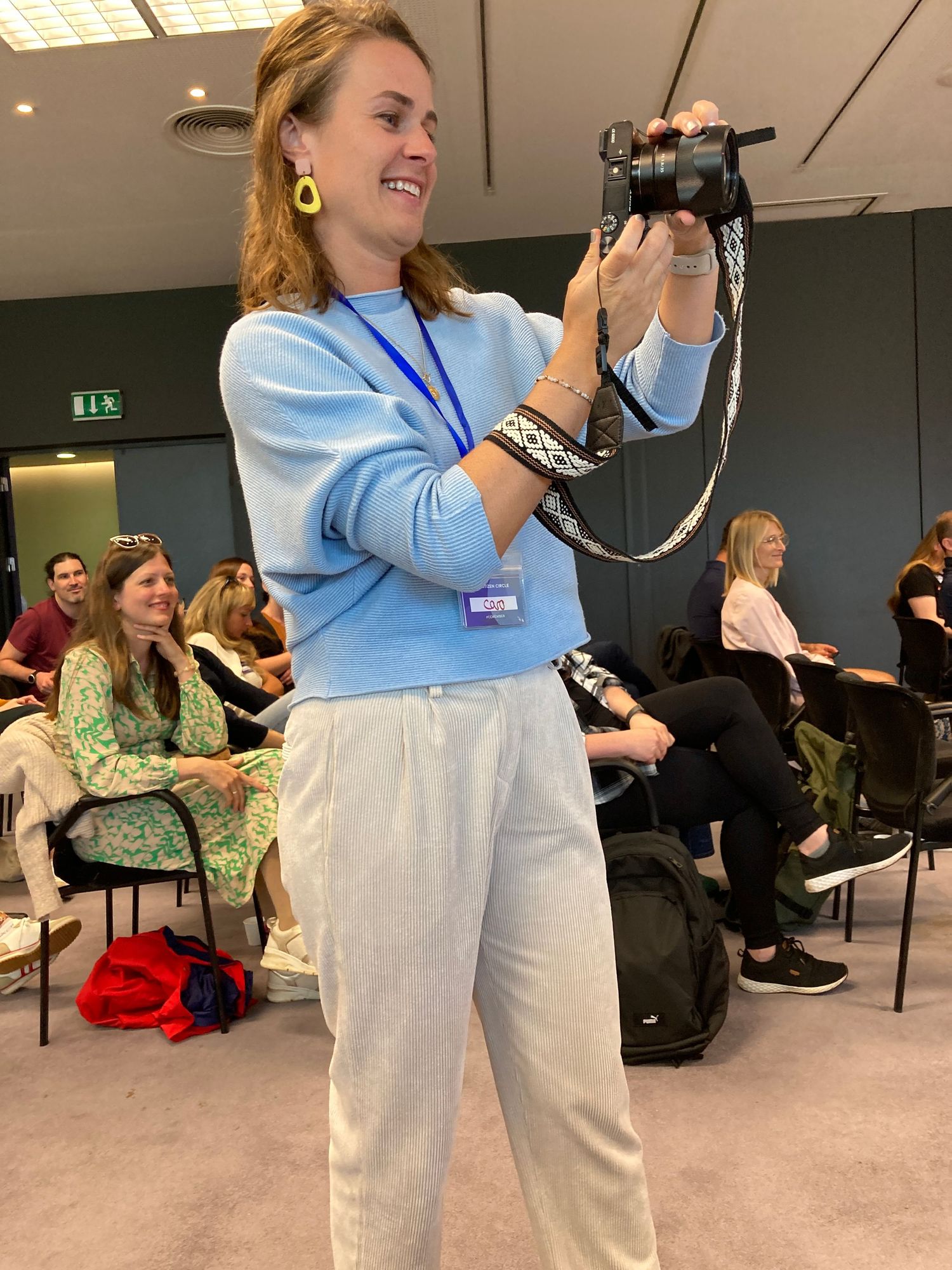 Woman in blue sweater smiling while photographing with camera at indoor conference event.