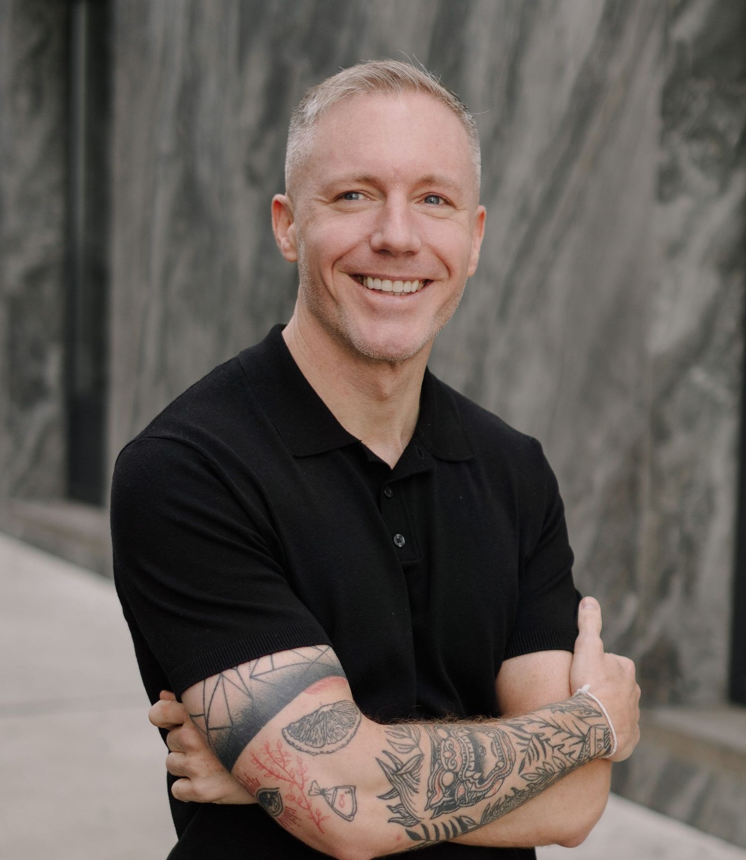 Smiling man with tattoos on arms crossed, wearing black polo shirt, standing outdoors against marble wall.