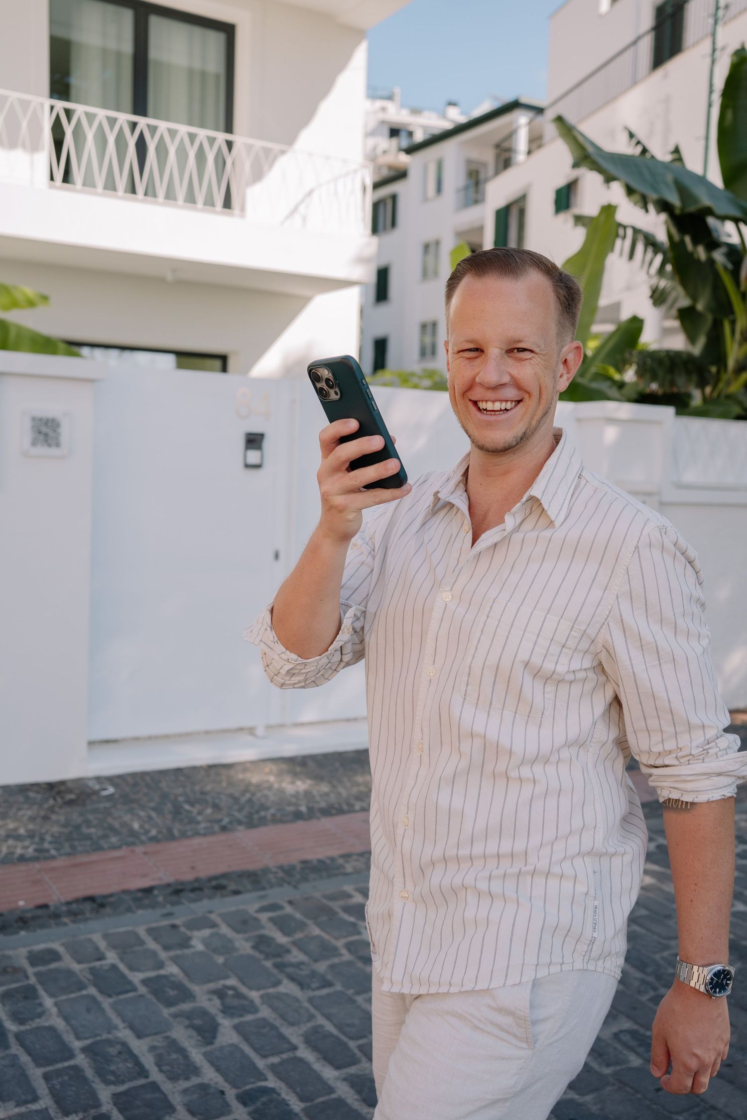 Smiling man in striped shirt holding smartphone outdoors near white Mediterranean buildings on sunny day.