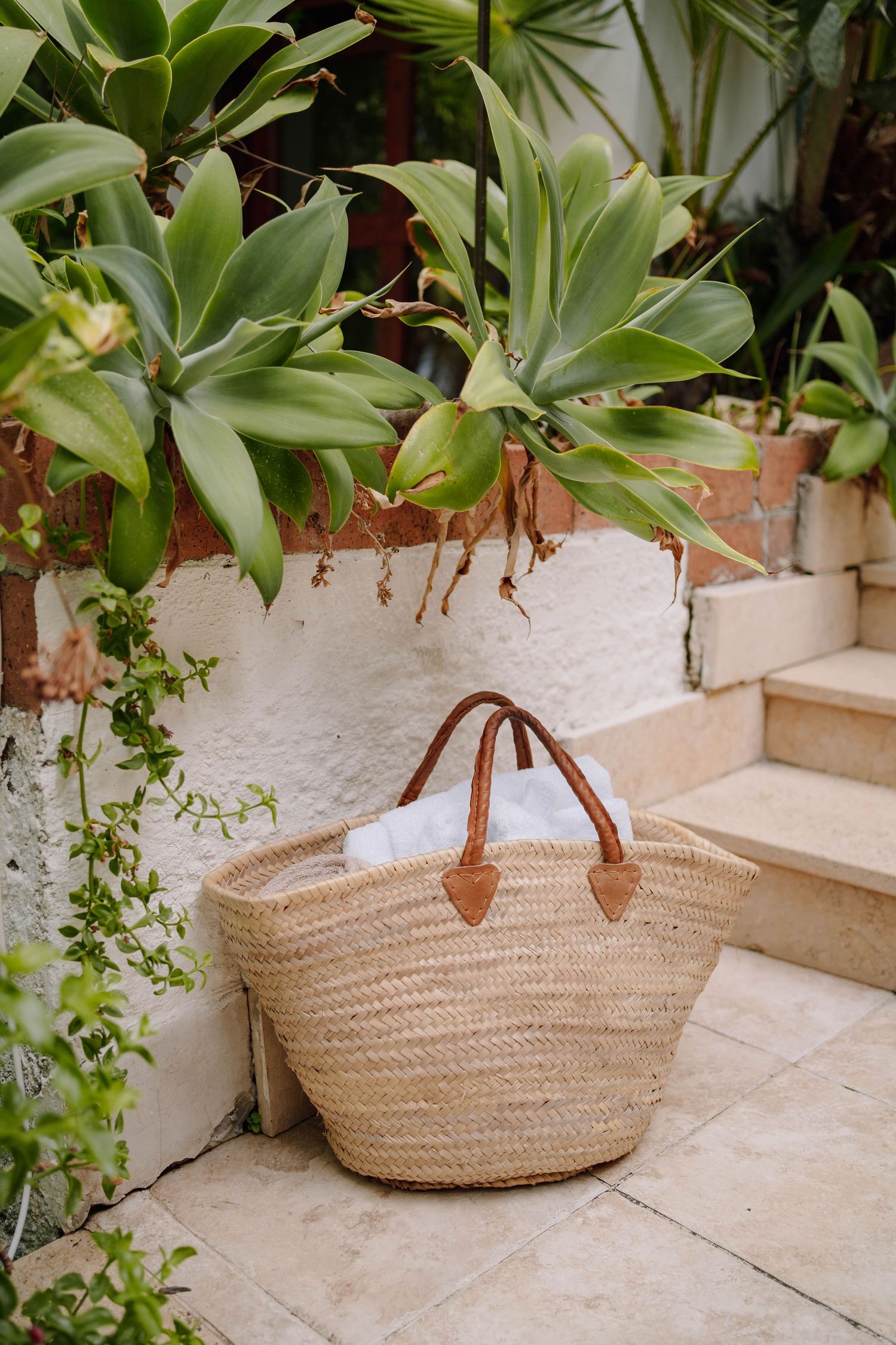 Woven straw tote bag with tan leather handles sits on a tiled patio beside a white wall, stone steps and lush green plants.