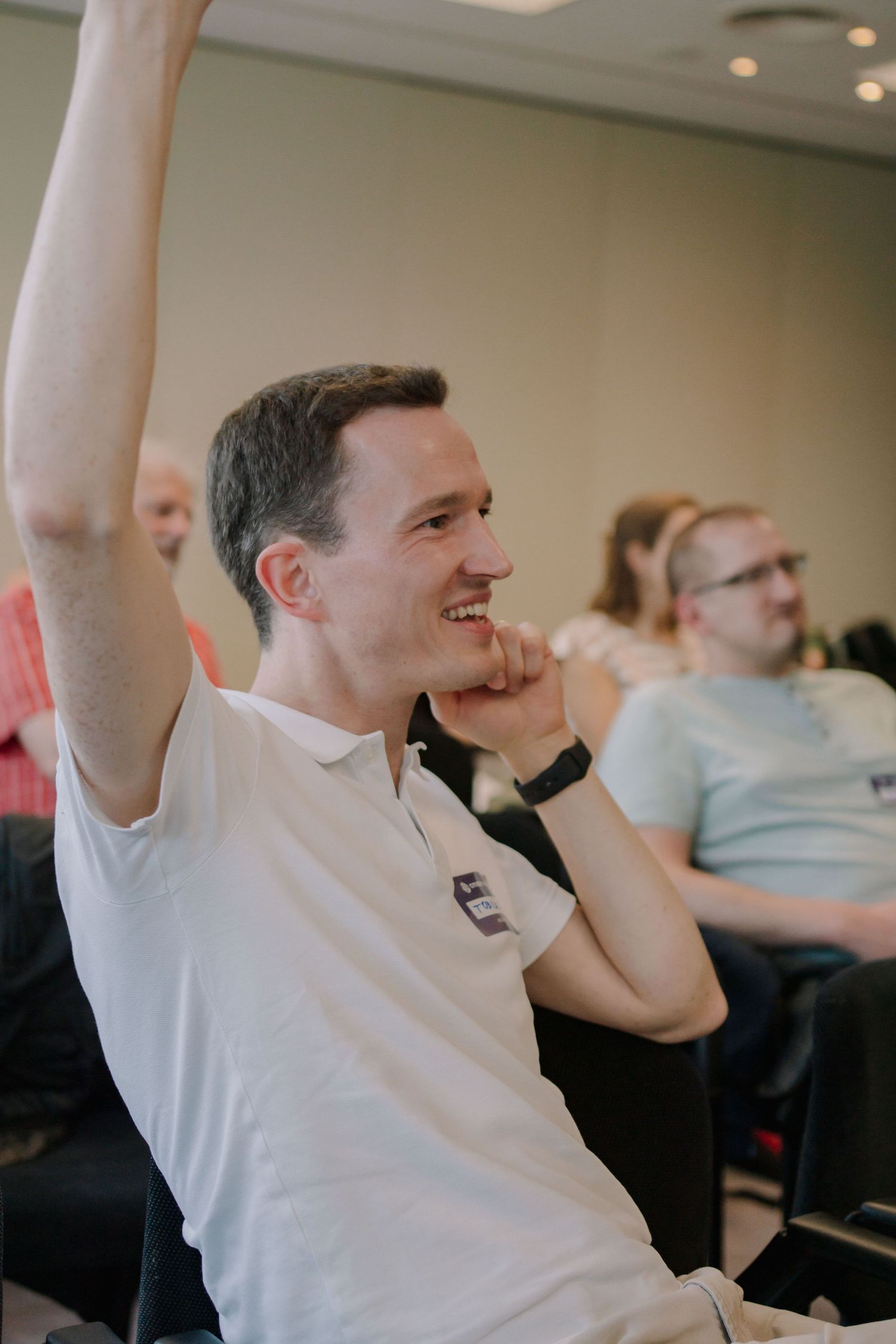 Smiling man in white polo shirt raising his hand during a conference or seminar event indoors.