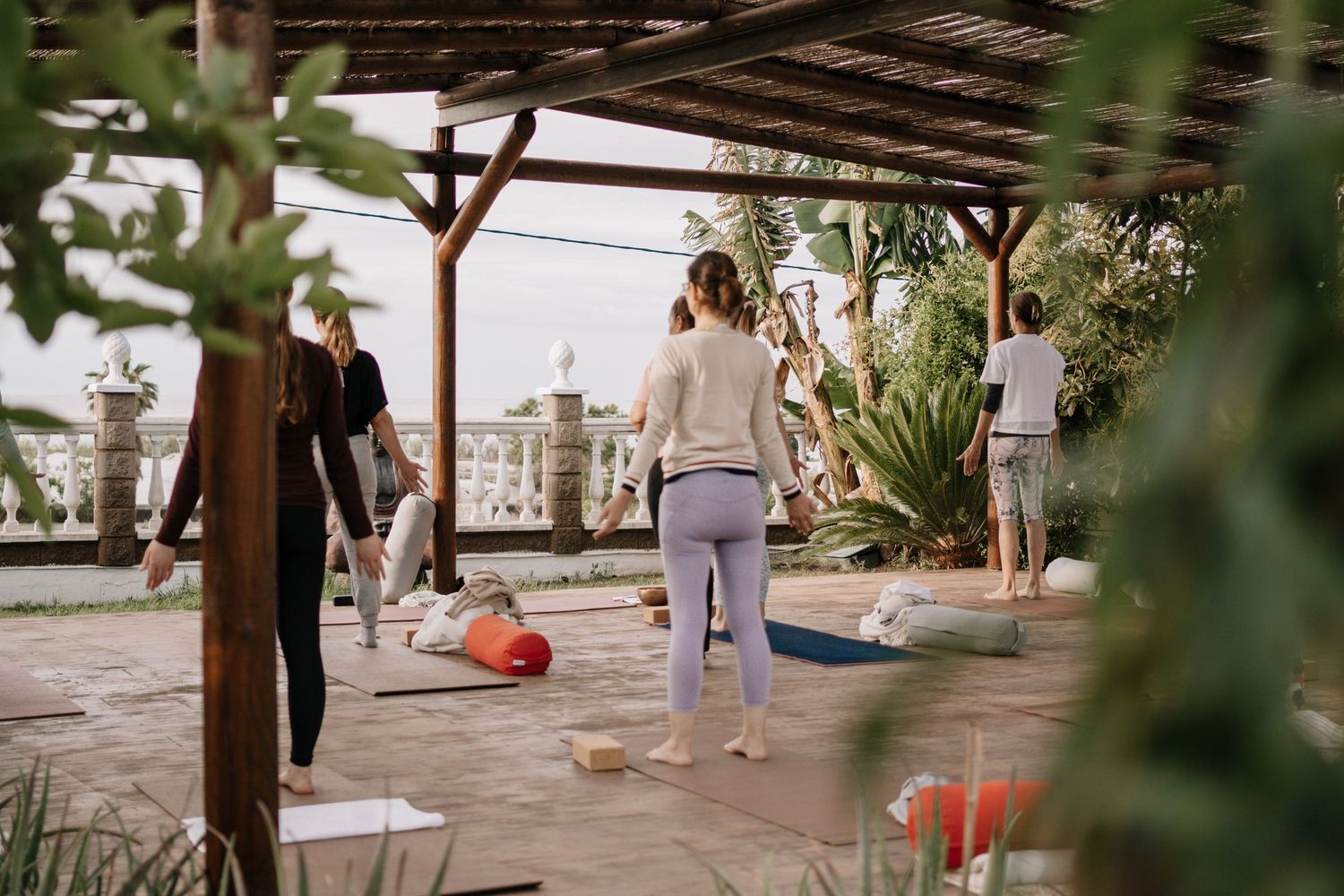 Group of people practicing yoga on an outdoor terrace under a wooden pergola surrounded by lush greenery.