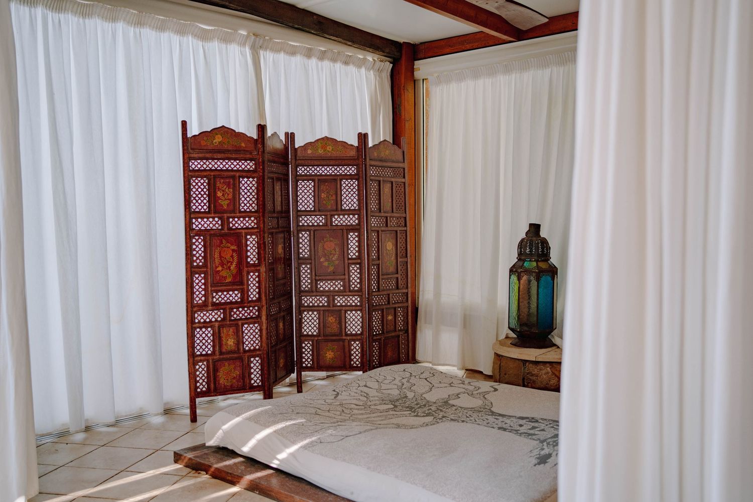 Carved wooden folding screen and a blue Moroccan lantern beside a daybed with white drapes in a bright, airy bohemian bedroom.