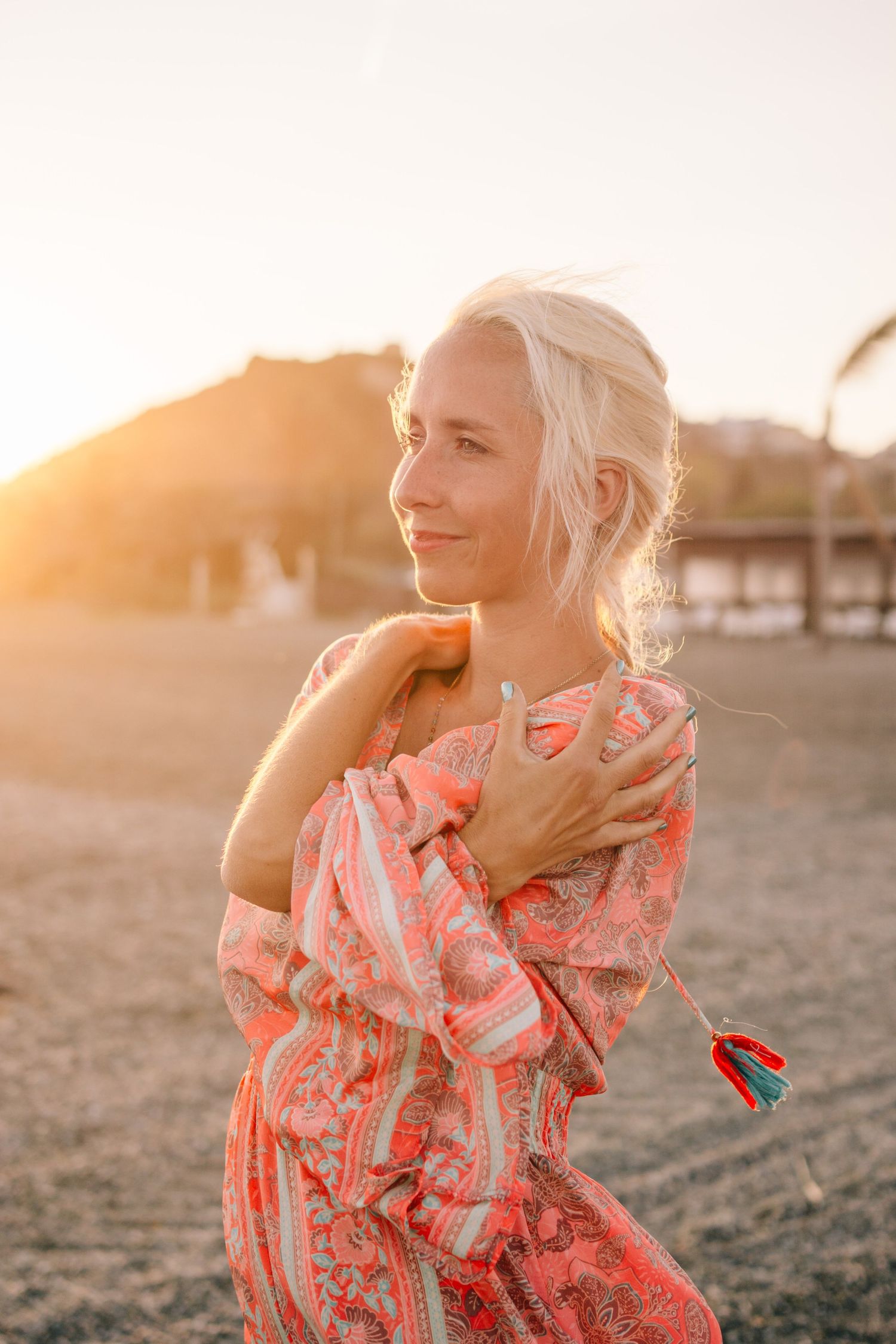 Blonde woman in pink floral dress posing on beach at golden sunset hour.