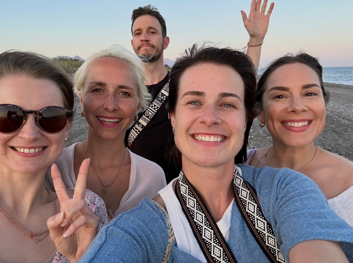 Group of five friends smiling for a beach selfie at sunset, with ocean waves visible in the background.