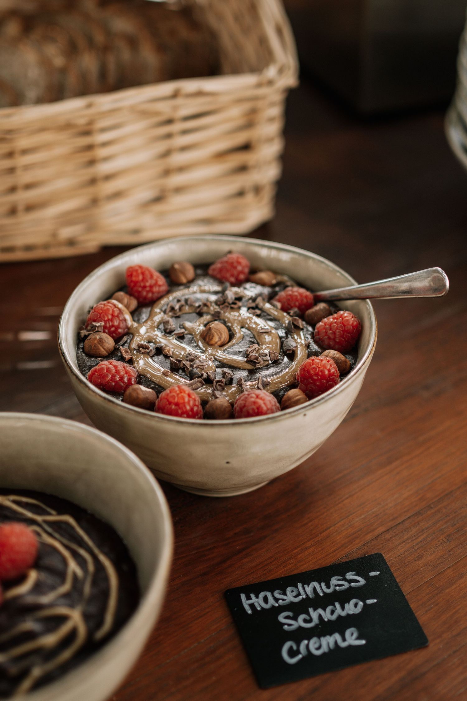 A ceramic bowl of hazelnut chocolate cream topped with raspberries and hazelnuts, labeled Haselnuss-Schoko-Creme.