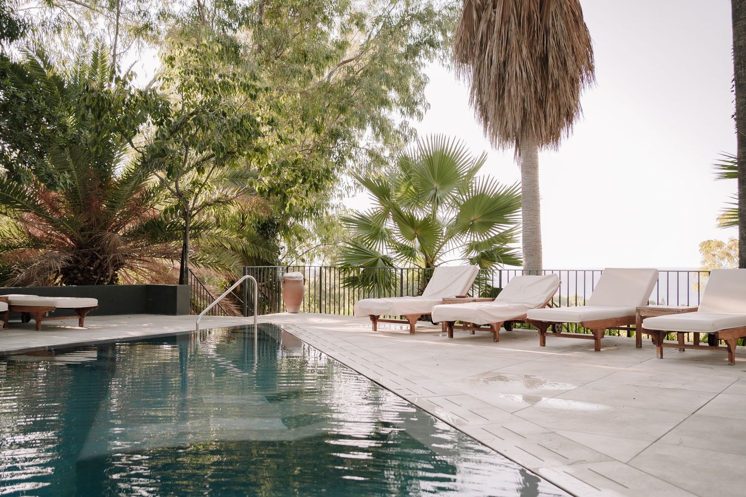 Outdoor swimming pool with wooden sun loungers on a stone terrace, surrounded by lush palm trees and tropical greenery.