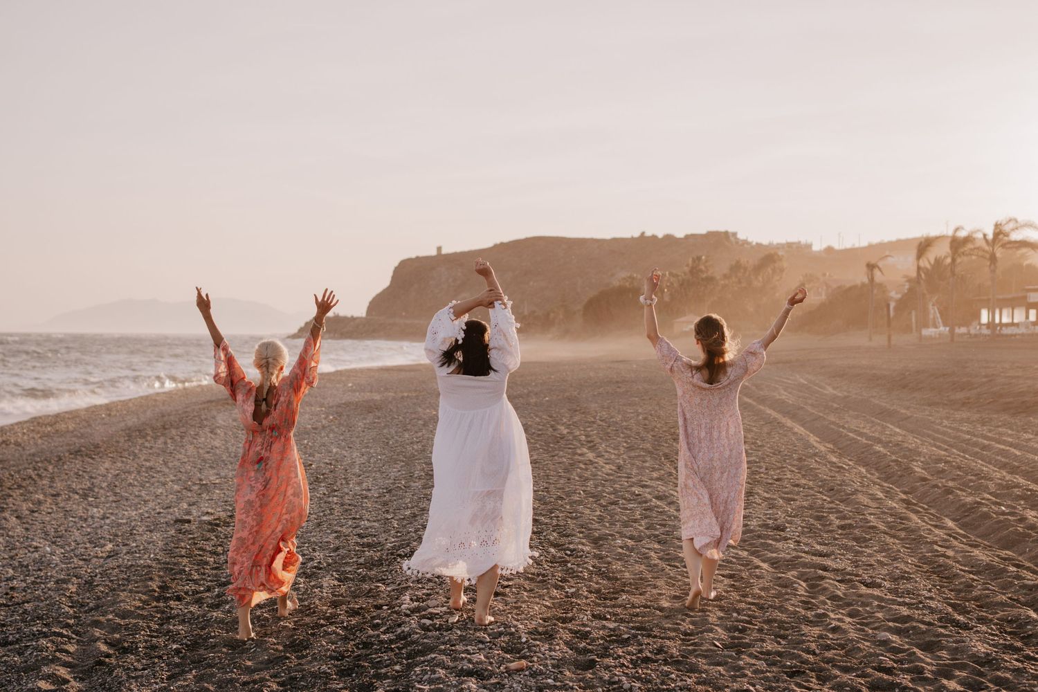 Three women in flowy dresses raising arms joyfully on sandy beach at golden sunset.