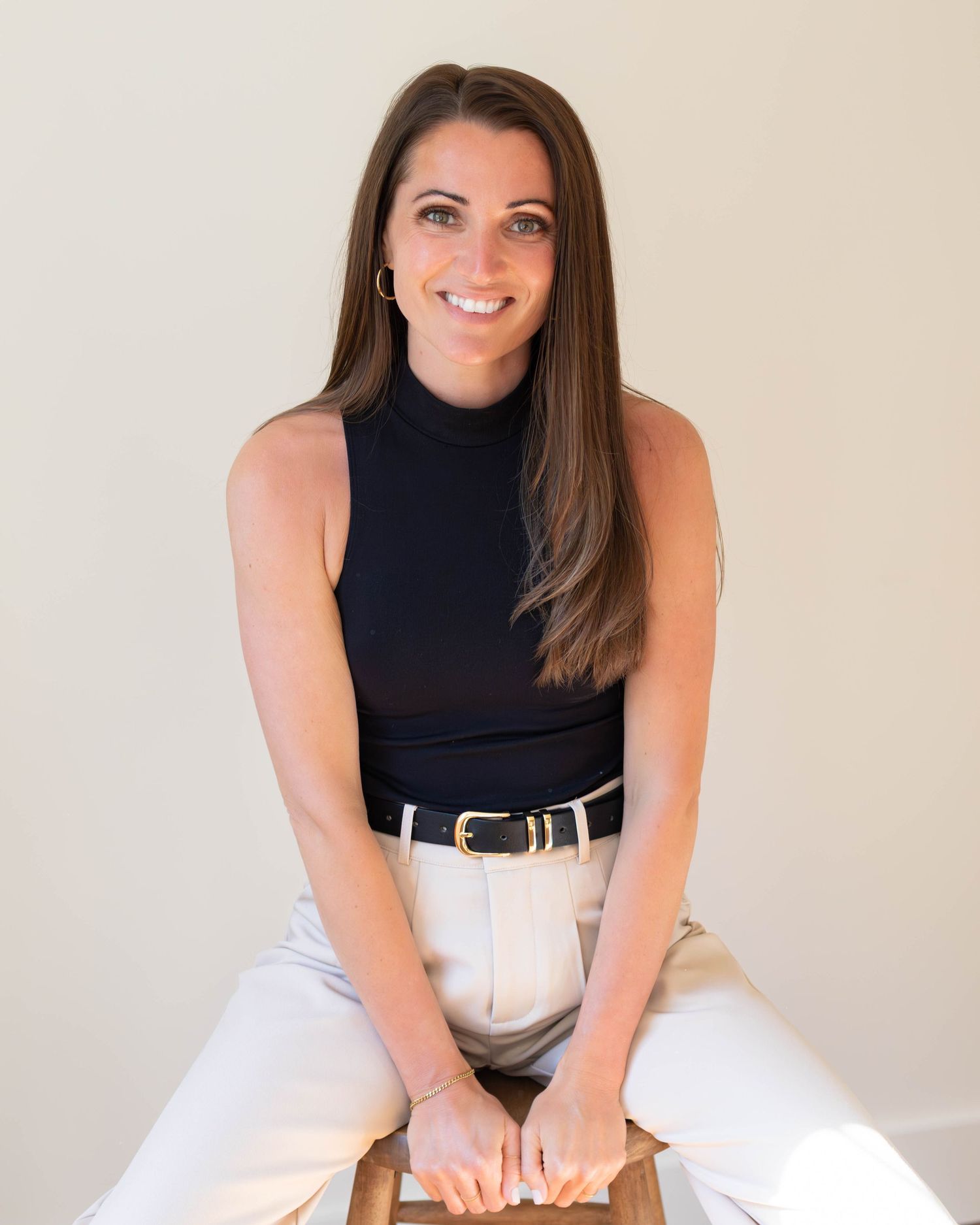A smiling brunette woman in a black turtleneck and white pants sits confidently on a stool against a neutral background.