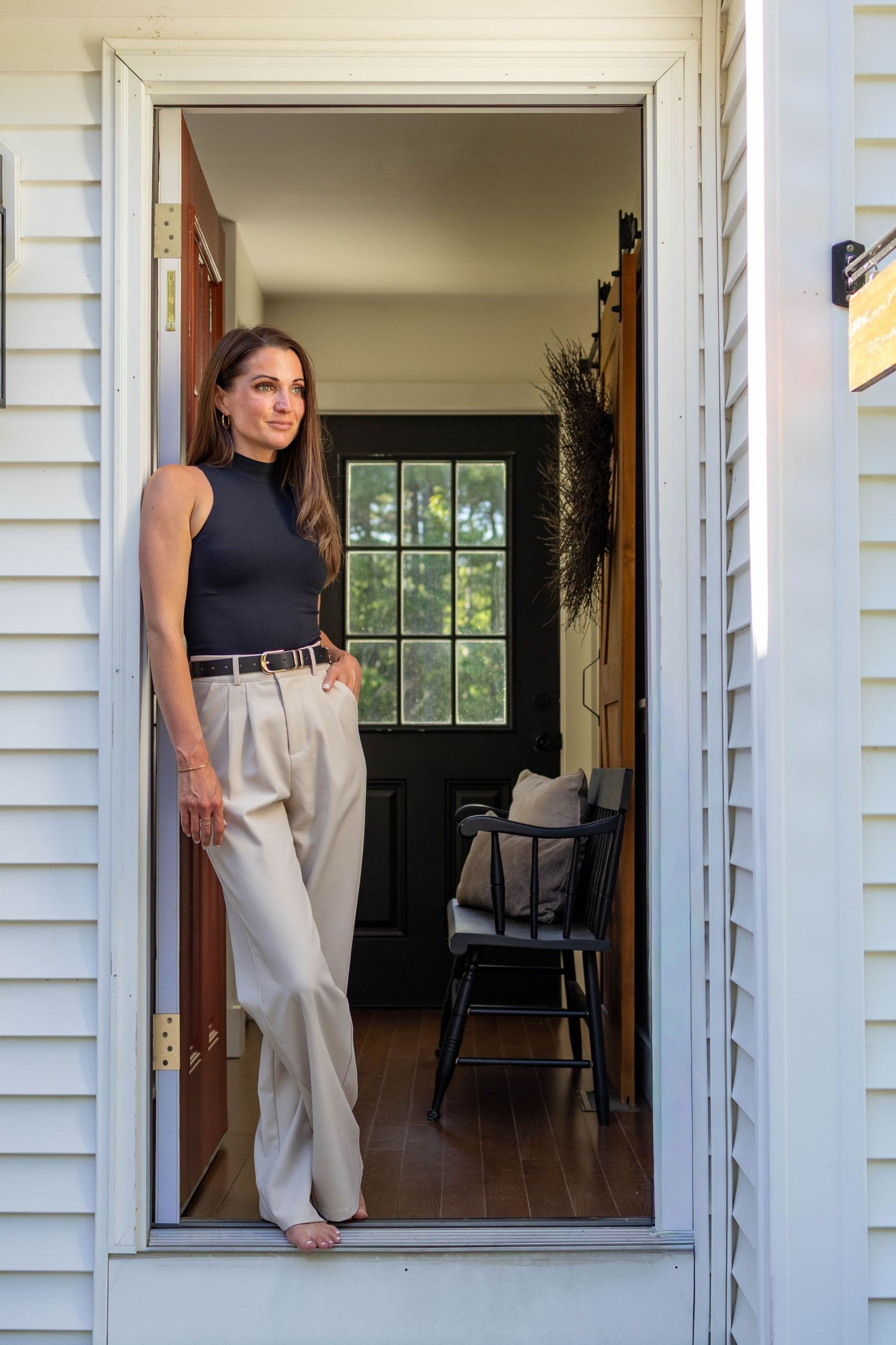 Woman in black sleeveless top and beige trousers standing in an open doorway of a white clapboard home.