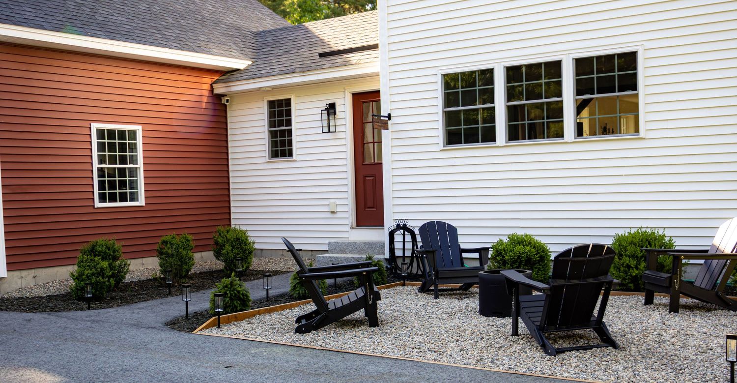 Charming home exterior with red and white siding, dark Adirondack chairs, gravel patio, and manicured shrubs.