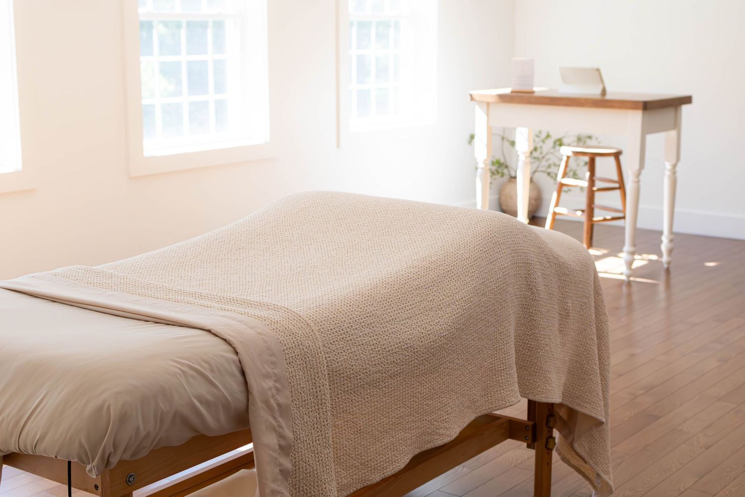 Serene massage therapy room with a draped treatment table, hardwood floors, bright windows, and a wooden desk in the background.