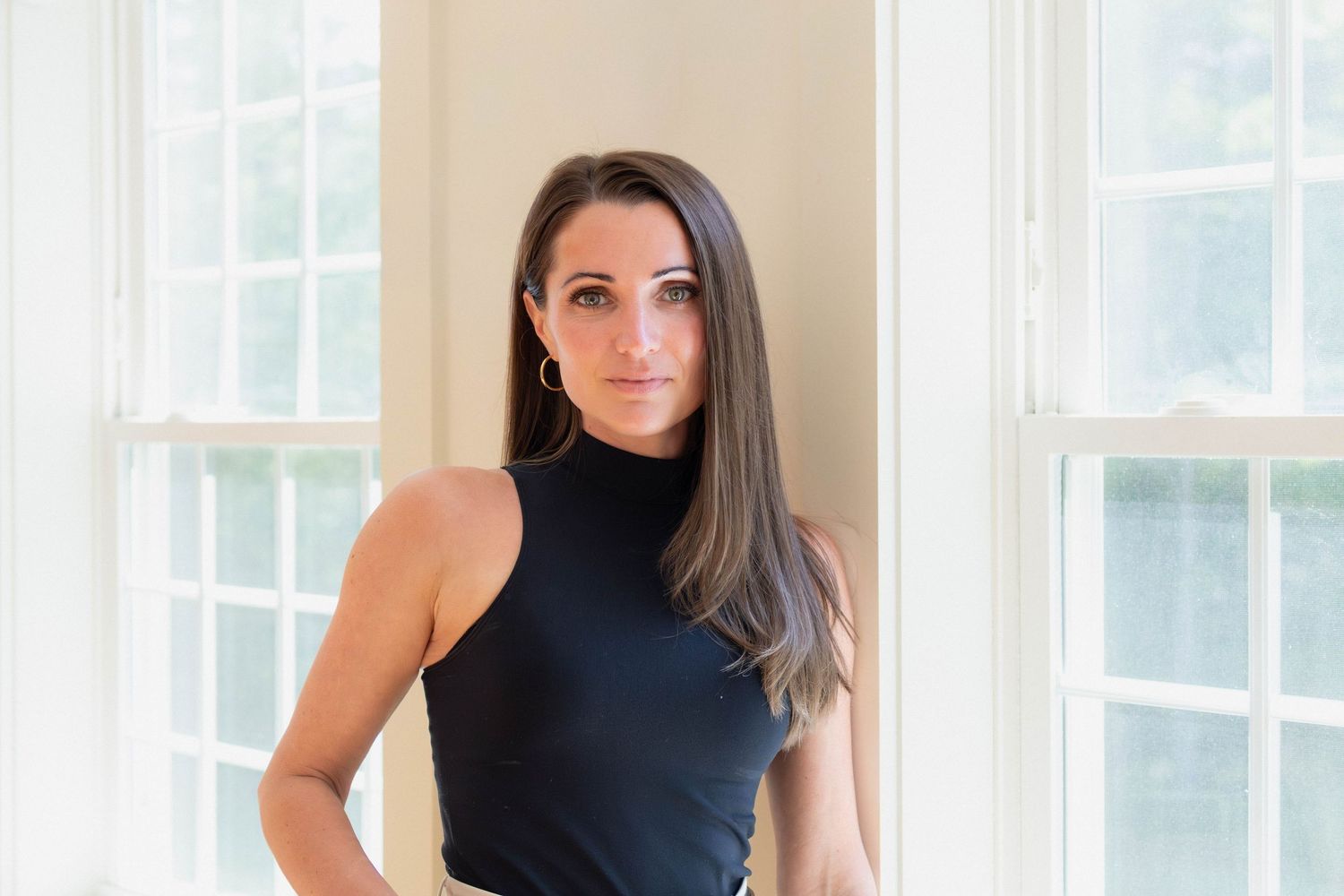 Woman in black sleeveless top standing between two windows in a bright room with natural light.
