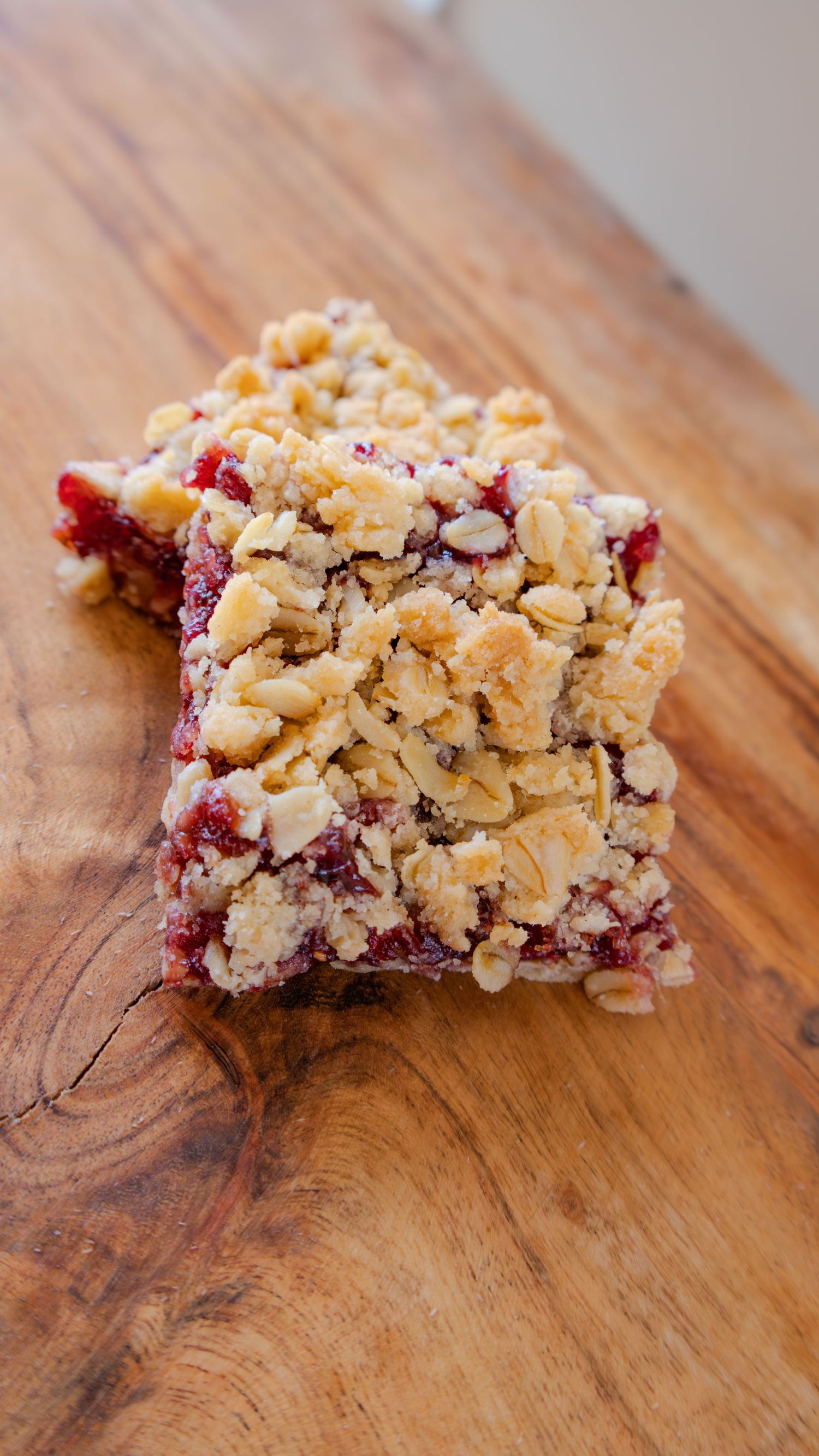 Two raspberry oat crumble bars stacked on a wooden cutting board, showing layers of jam and golden oat topping.