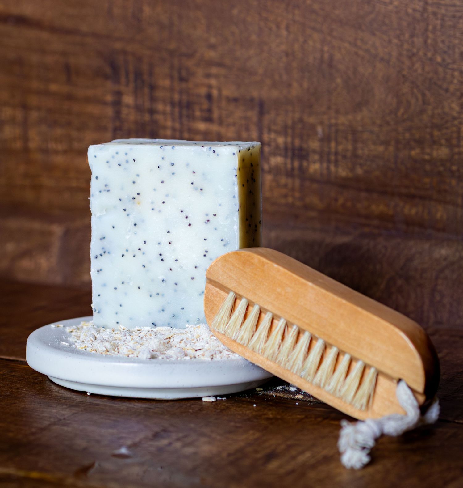 Handmade speckled soap bar on ceramic dish with wooden nail brush on rustic wood surface.