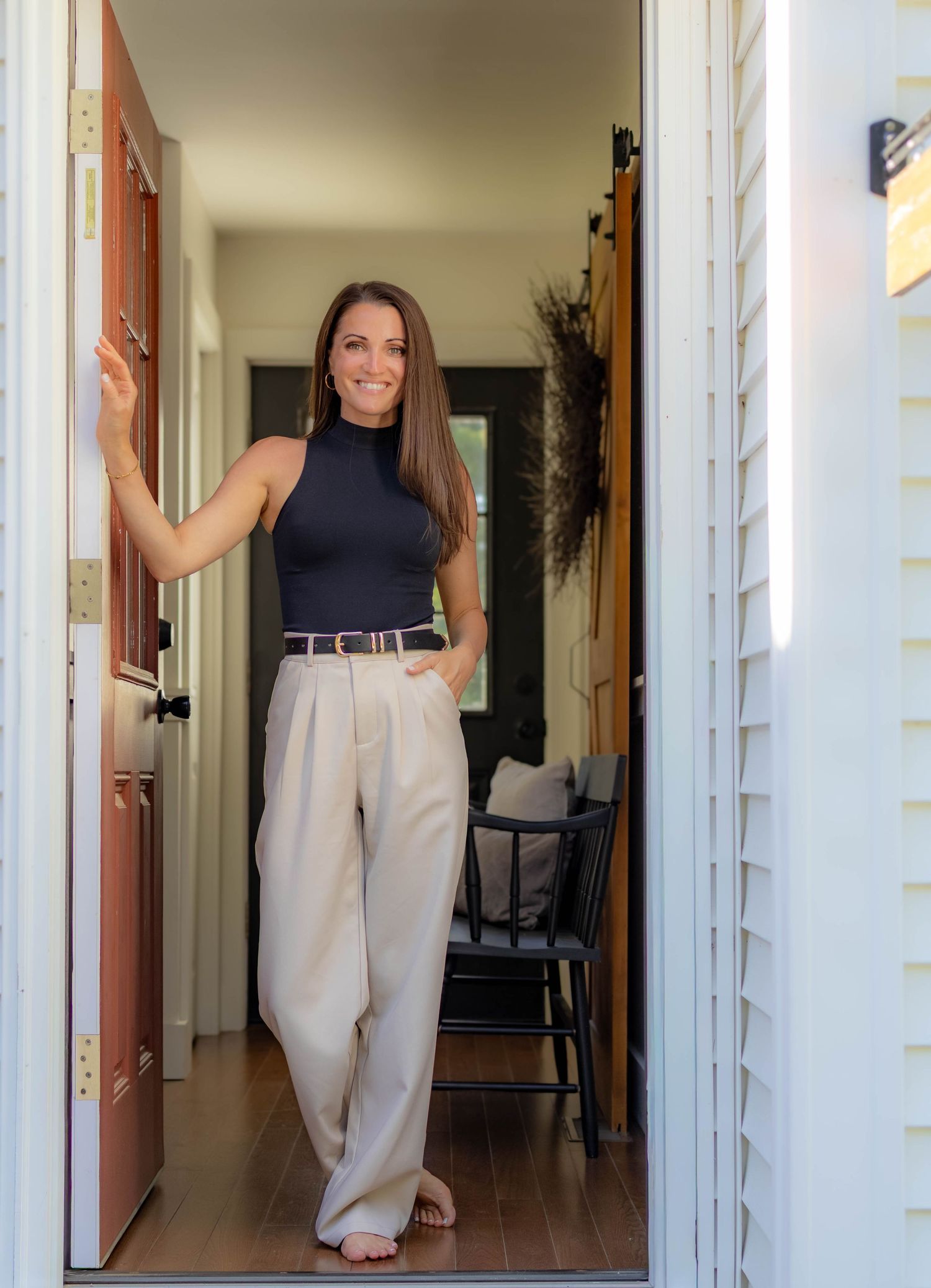 Smiling woman in black sleeveless top and beige wide-leg pants standing in an open doorway of a modern home.