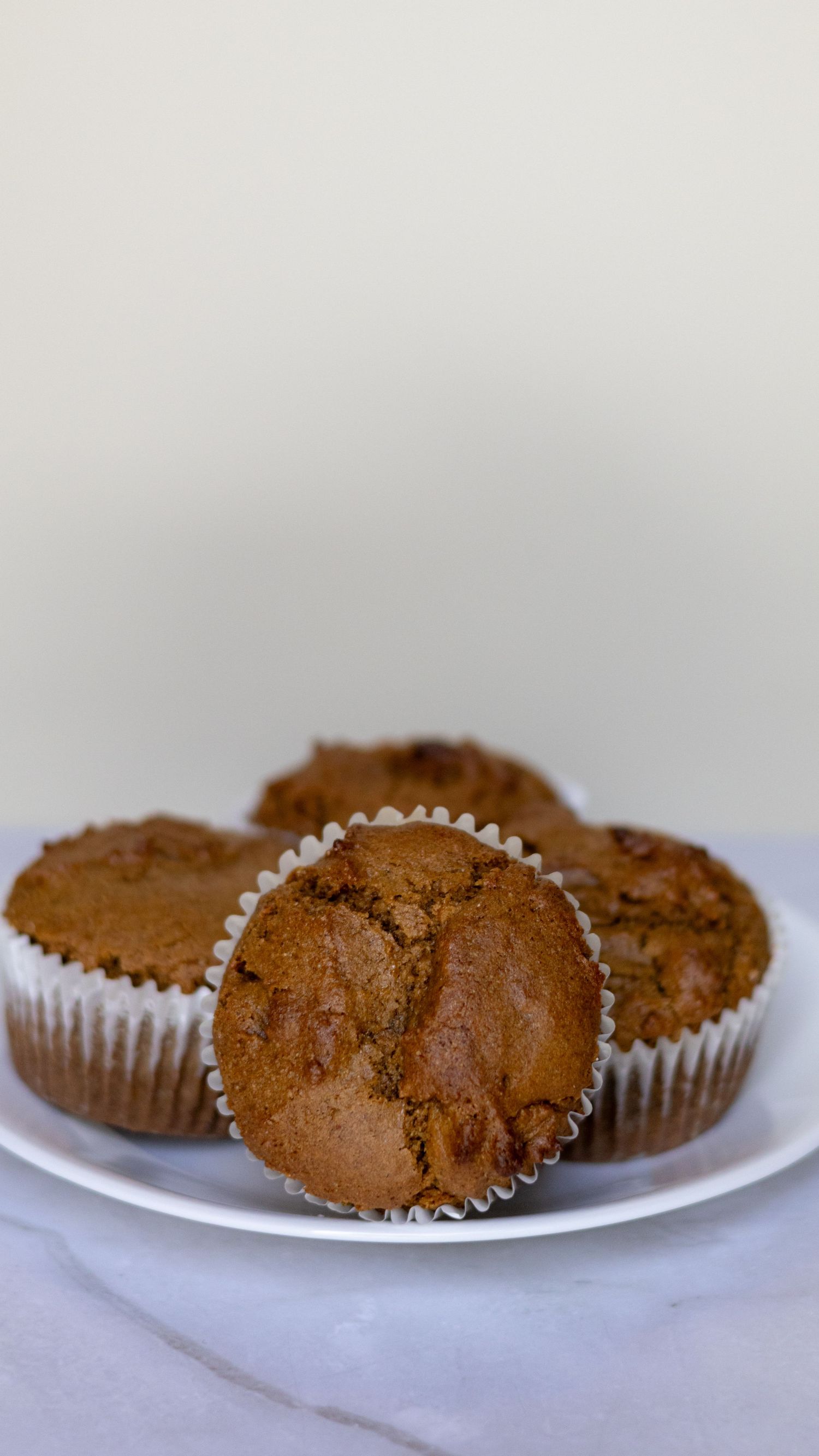 Four golden-brown muffins in white paper liners arranged on a white plate against a light background.