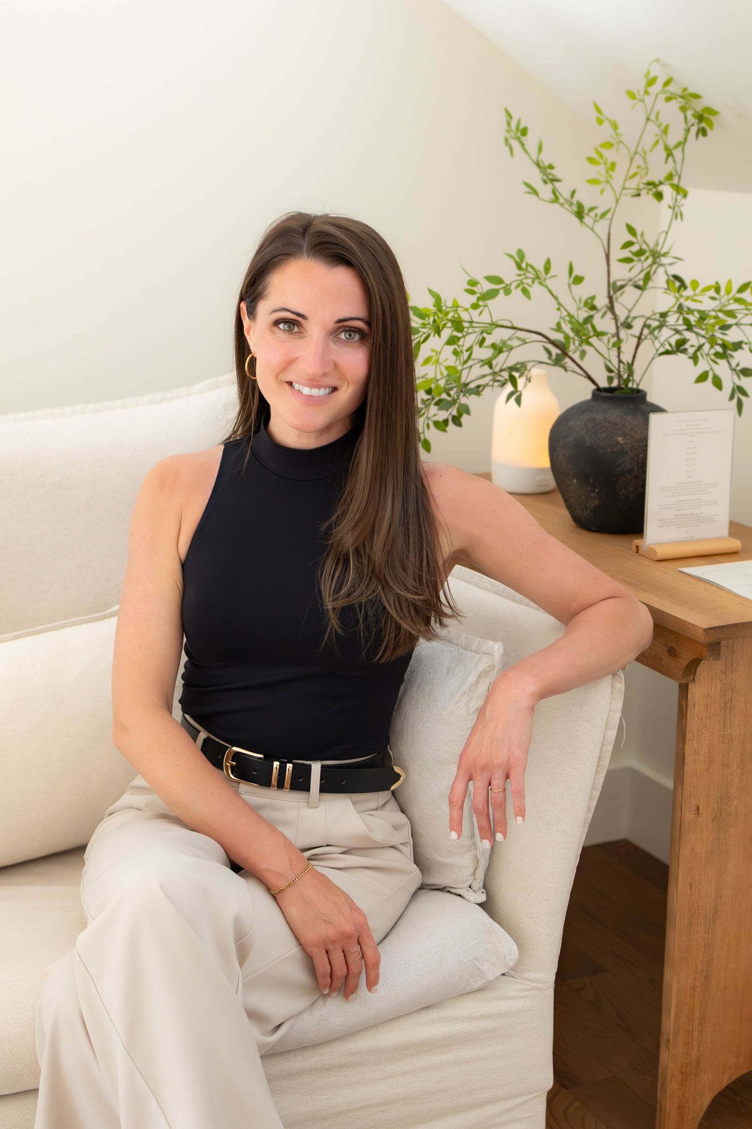 A smiling brunette woman in a black sleeveless top sits relaxed on a cream sofa in a bright, modern living space.