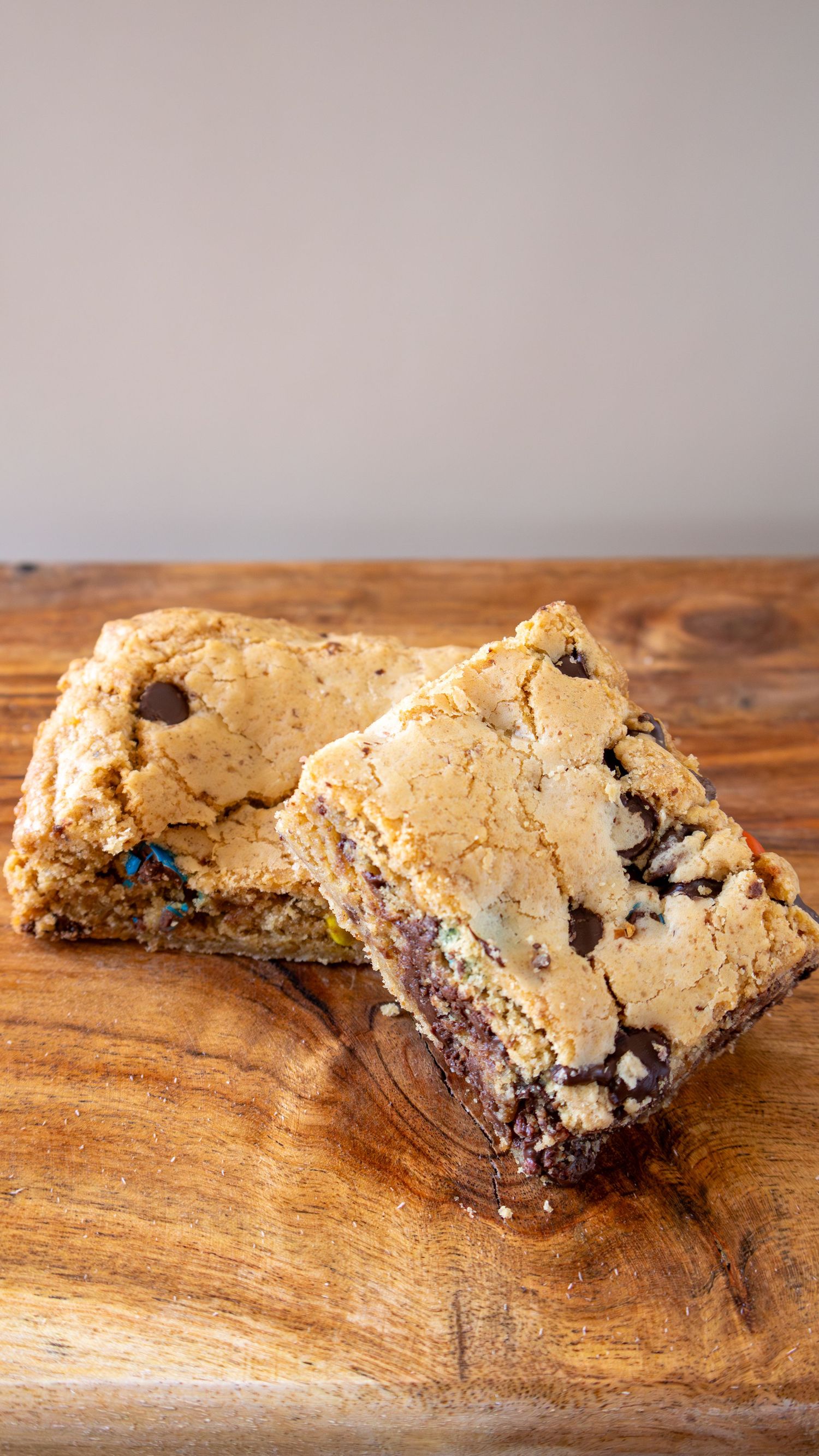 Two golden chocolate chip cookie bars with colorful candy pieces stacked on a rustic wooden cutting board.