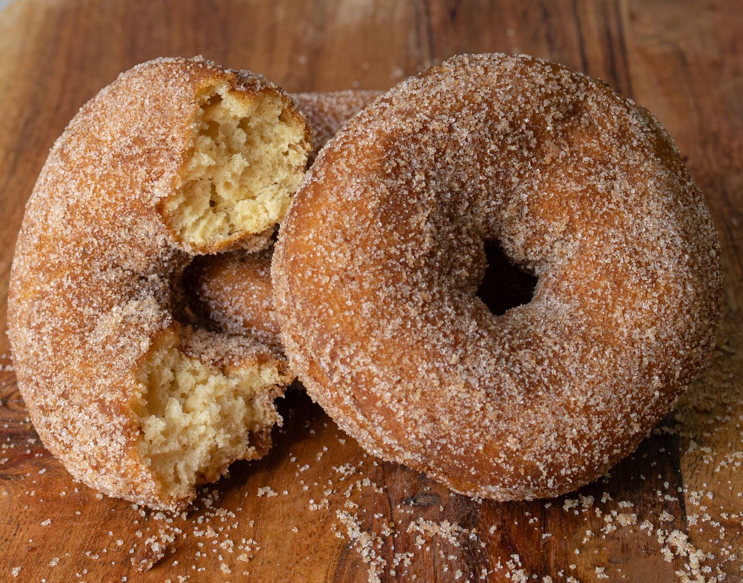 Two cinnamon sugar-coated donuts on a wooden board, one with a bite taken out revealing soft, fluffy interior.