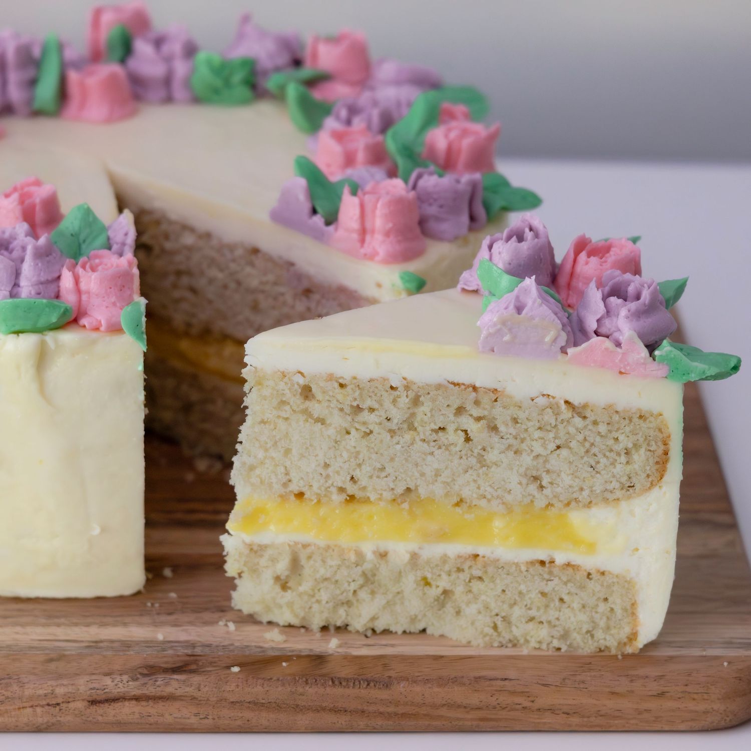 A two-layer vanilla cake with lemon curd filling, white frosting, and pink and purple buttercream flowers on a wooden board.