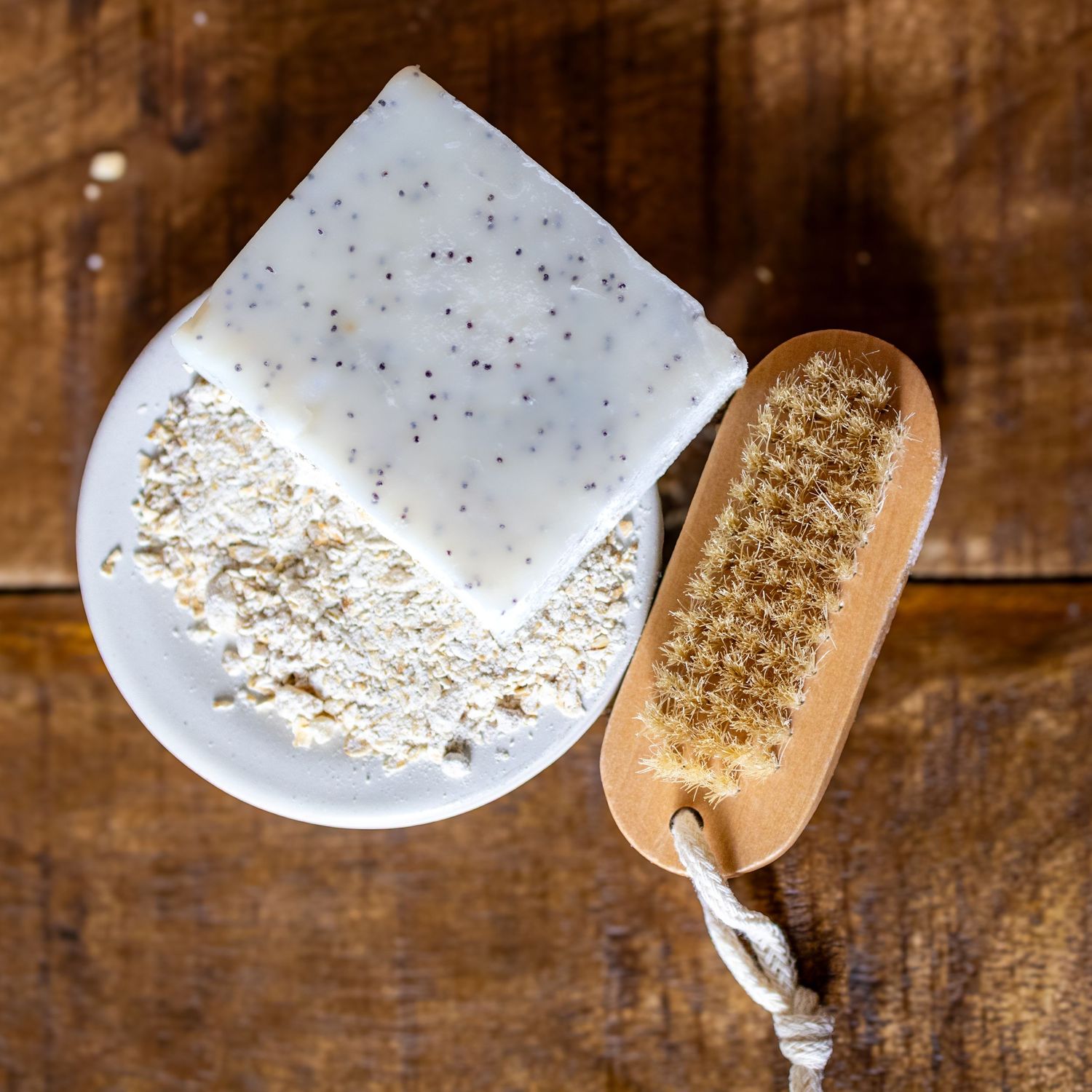 Handmade speckled soap bar with oat powder and a wooden nail brush on a rustic wood background.