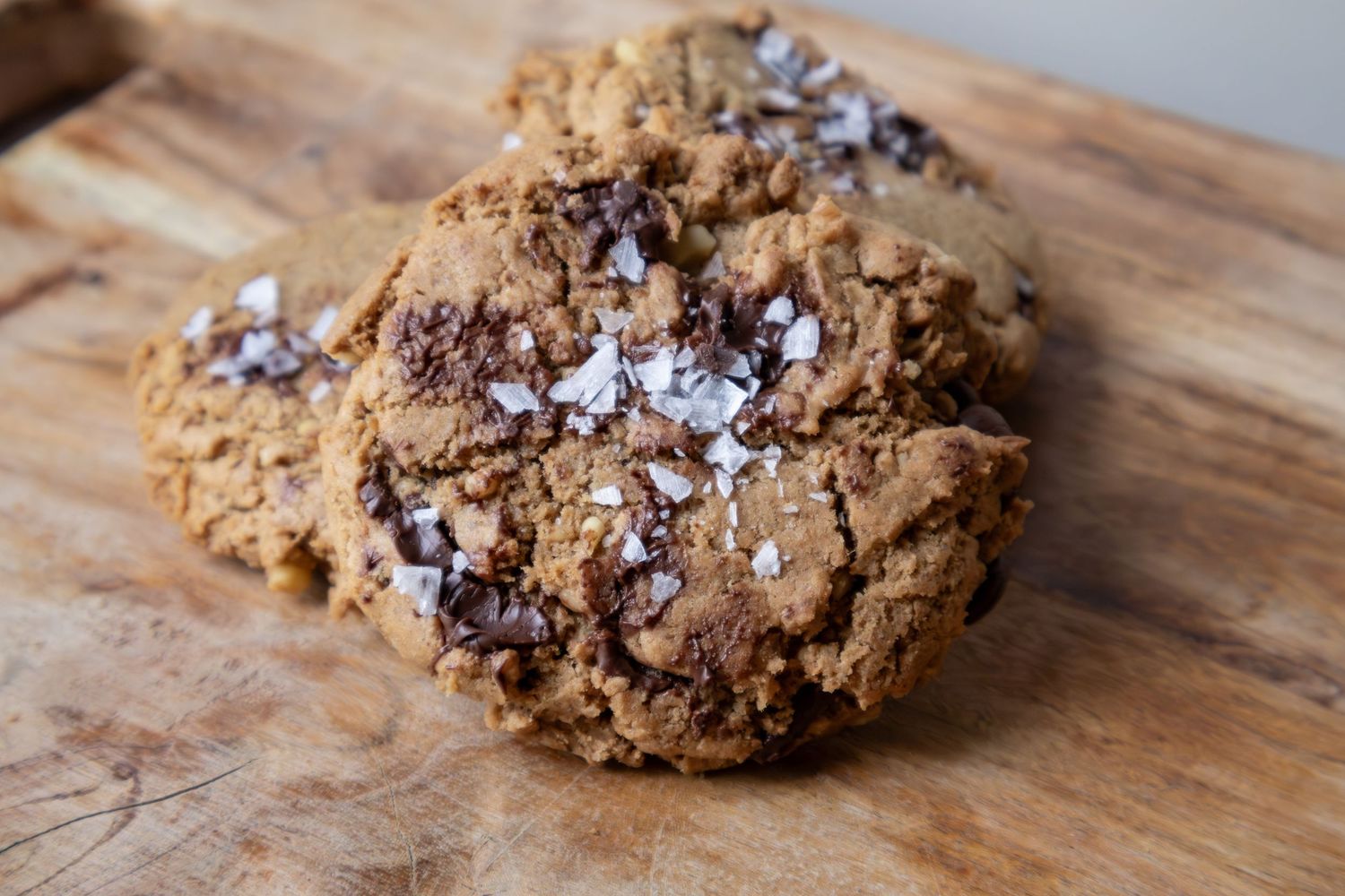 A large chocolate chip cookie topped with flaky sea salt on a wooden cutting board.