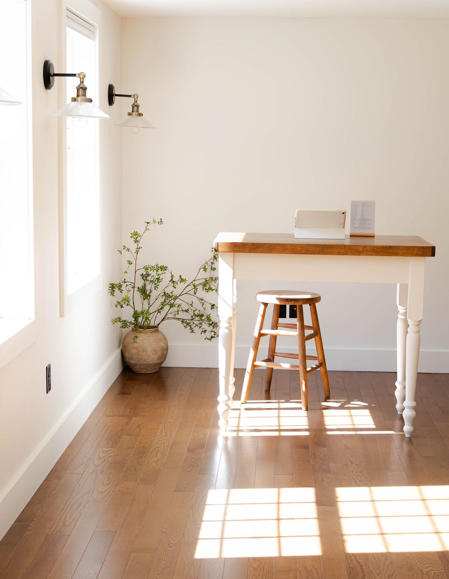 Minimalist home office with white farmhouse desk, wooden stool, potted plant, and warm sunlight on hardwood floors.