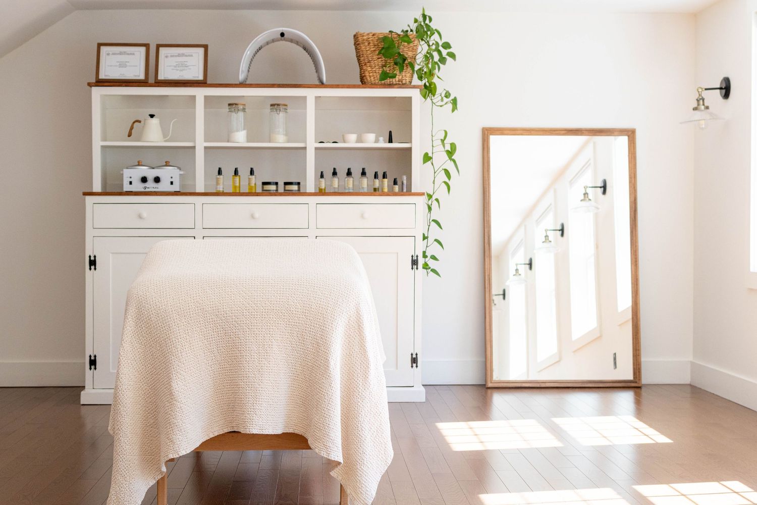 Bright spa treatment room with white storage cabinet, massage table draped in linen, large mirror, and natural light on hardwood floors.