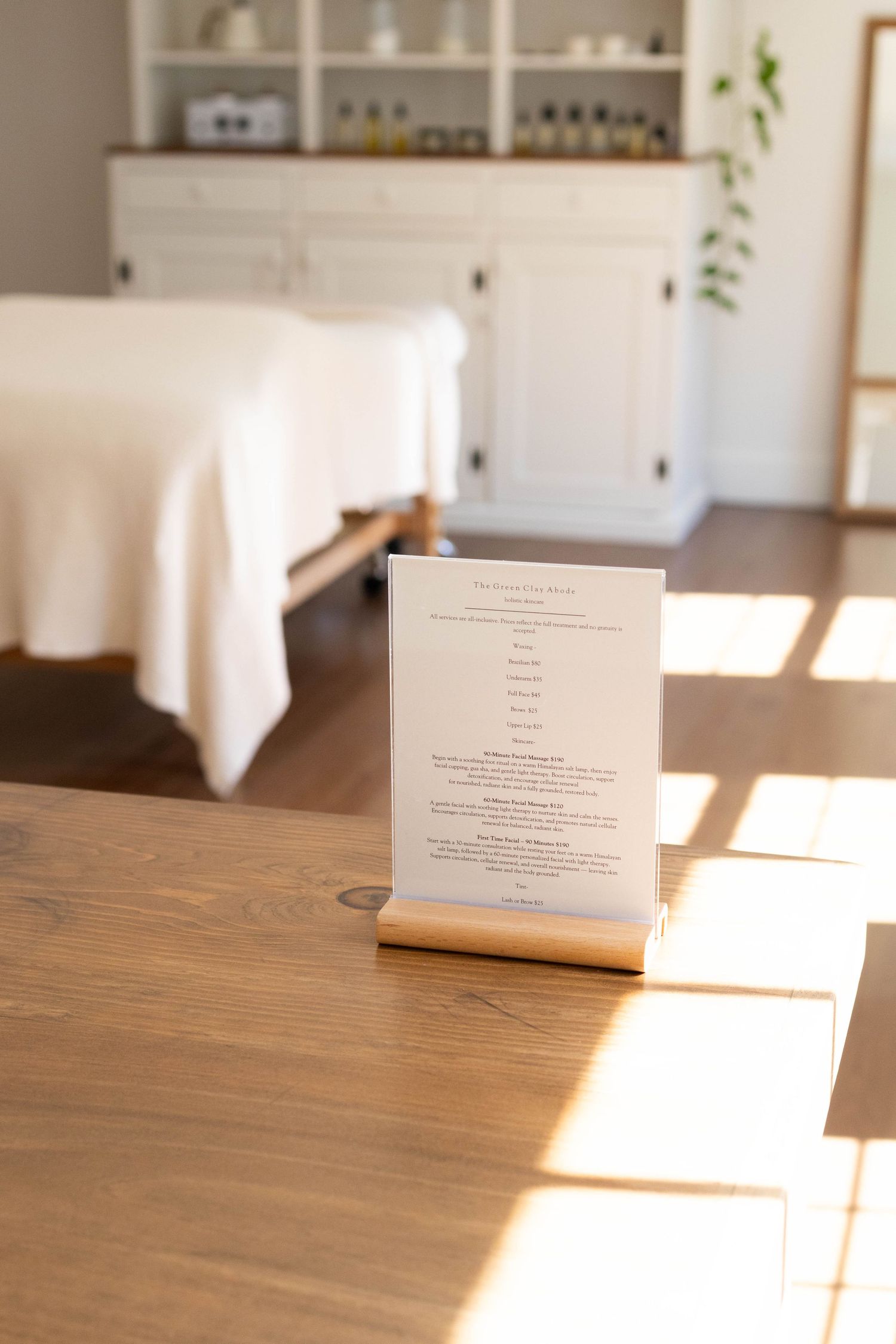 A menu card on a wooden table in a sunlit spa room with a massage table and white cabinet in the background.