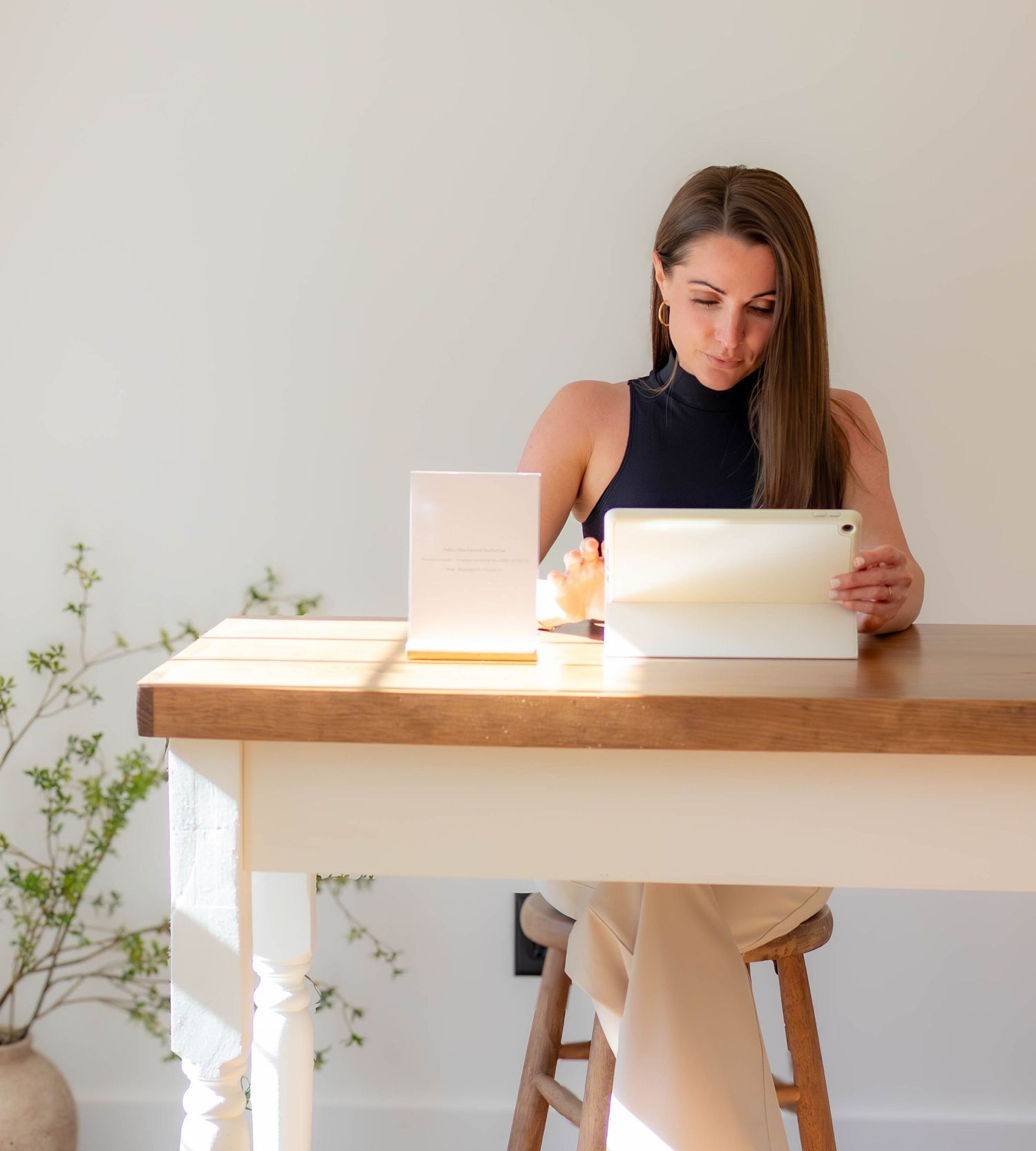 Esthetician sitting at a wooden desk using a tablet, with books and stationery in a bright, minimalist home office.