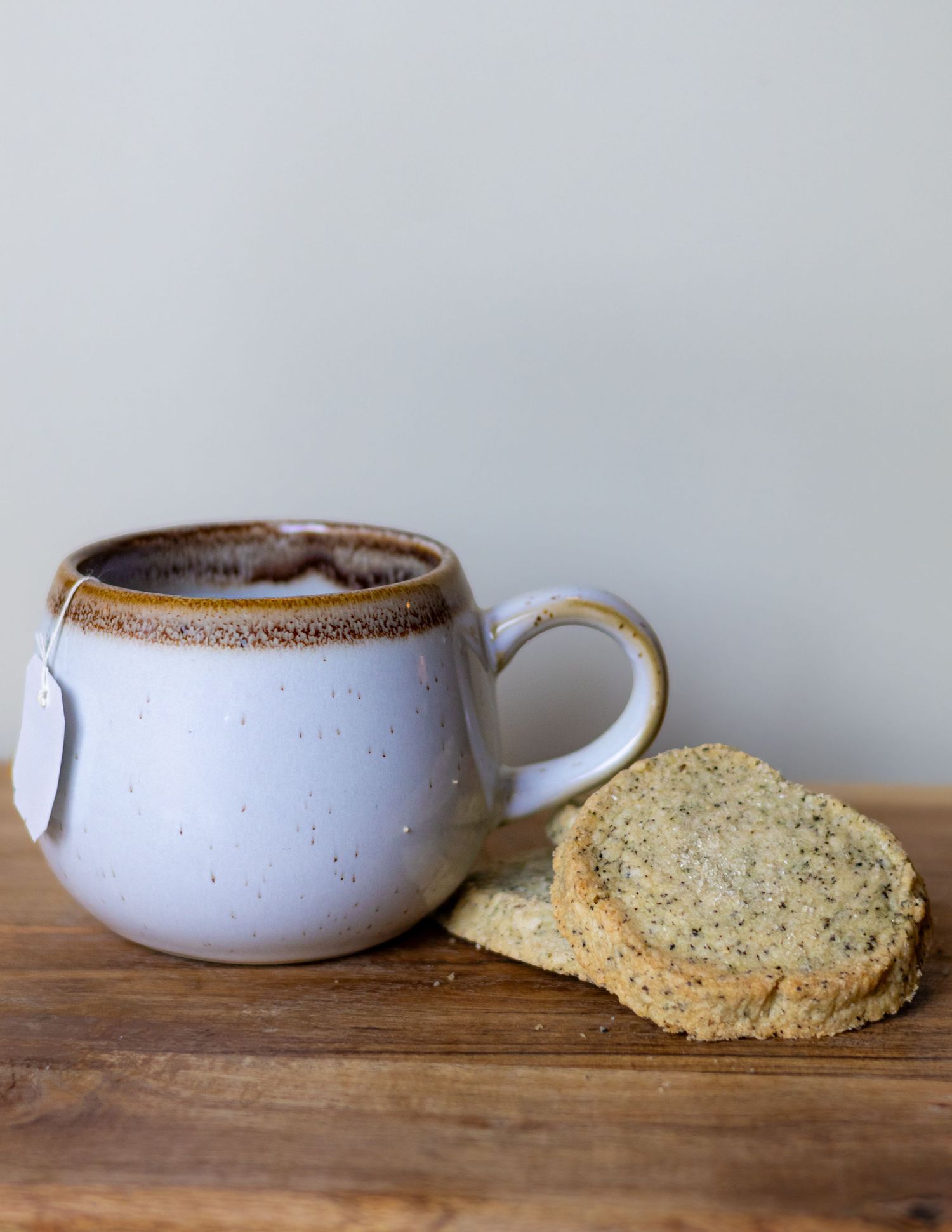 Rustic speckled ceramic mug with tea bag beside two round shortbread cookies on a wooden surface.