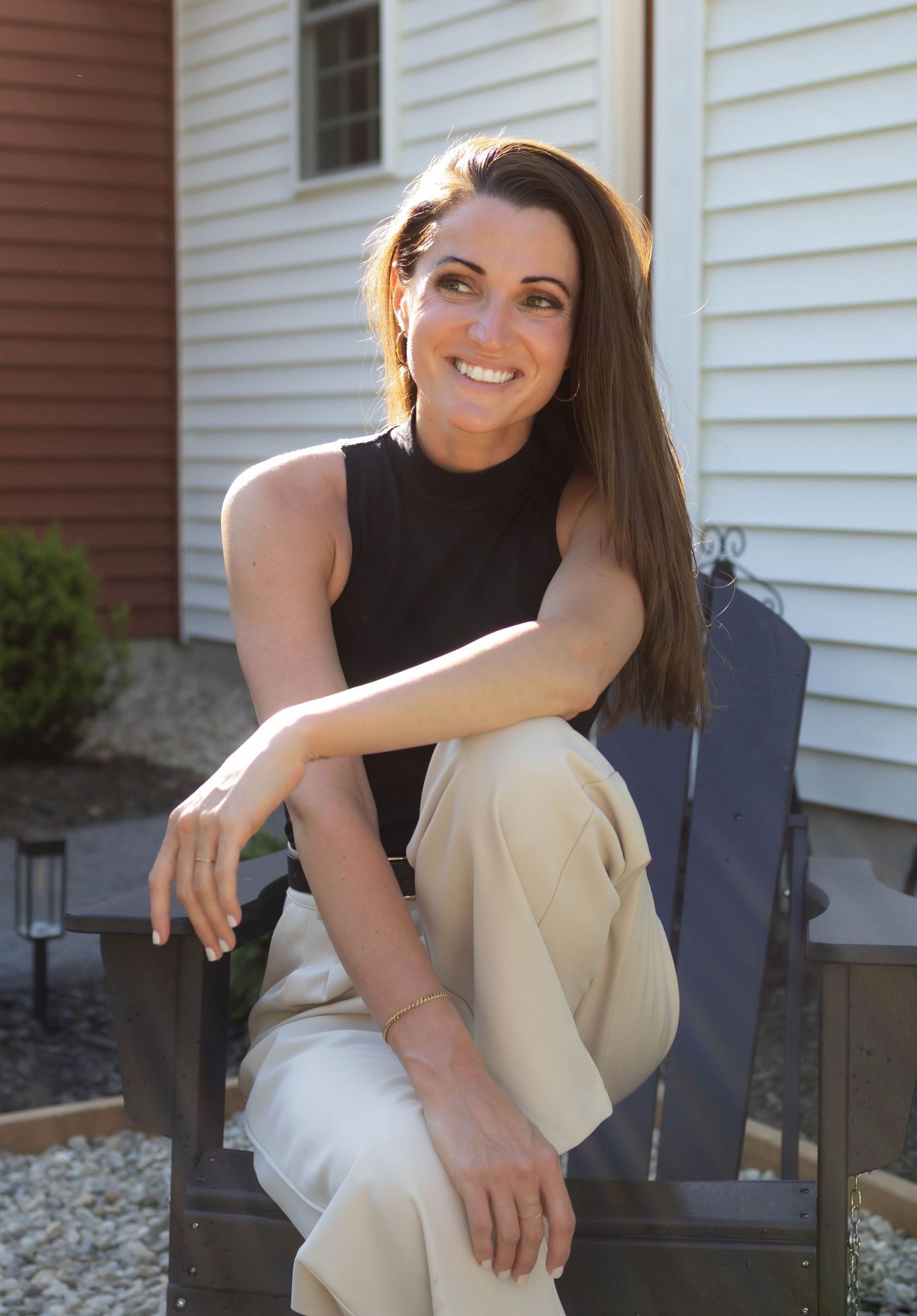 Smiling business owner woman in black sleeveless top and beige pants sitting outdoors near a house.