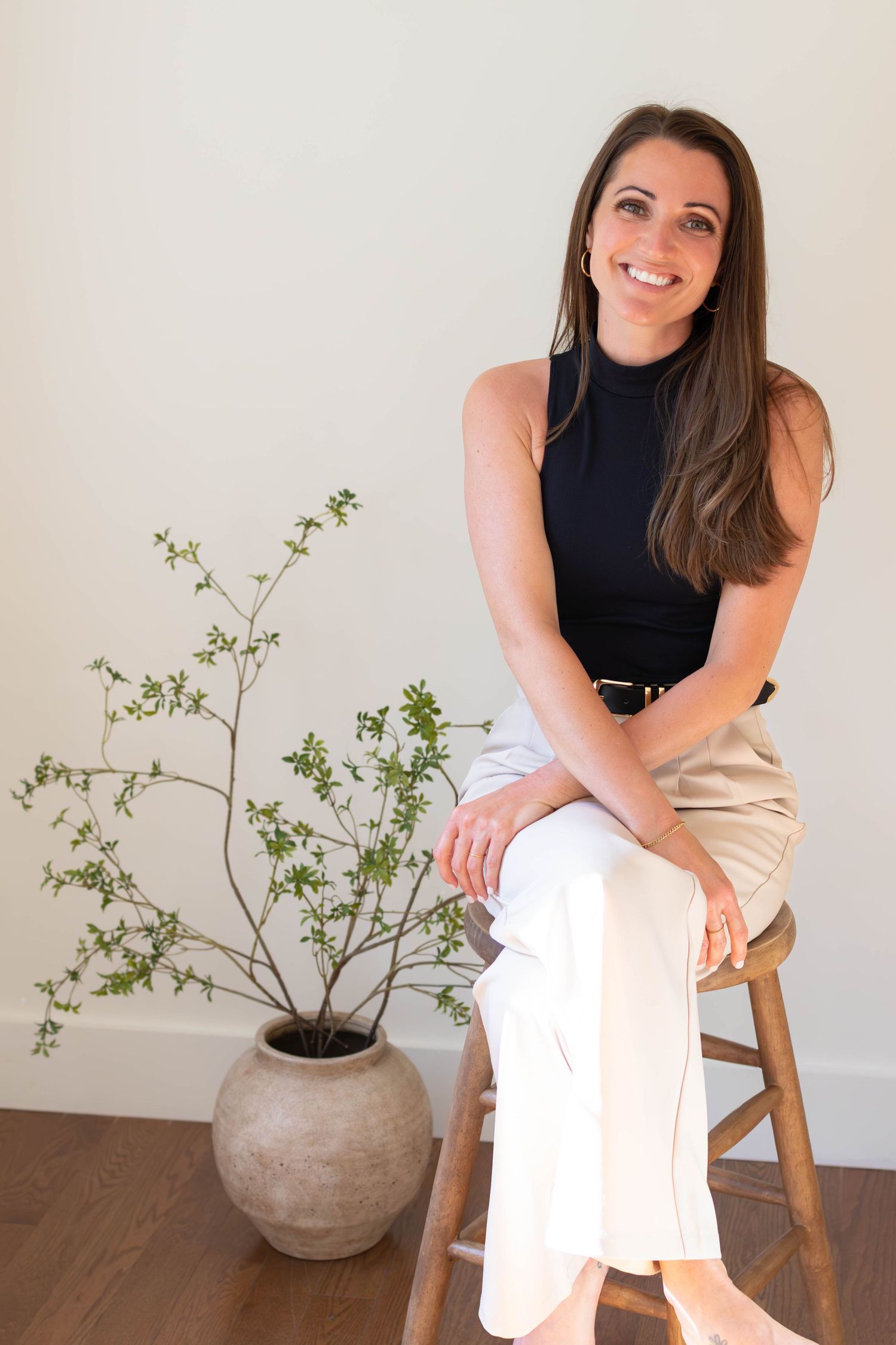 Smiling brunette woman in black top and white pants sitting on a wooden stool beside a potted plant.