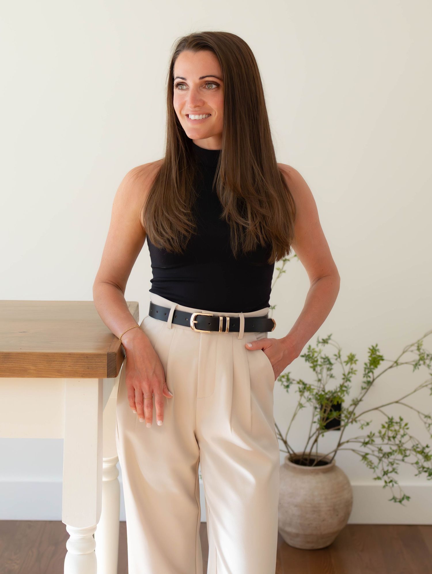 Woman in black sleeveless top and cream wide-leg trousers with belt, smiling and posing in a bright modern interior.