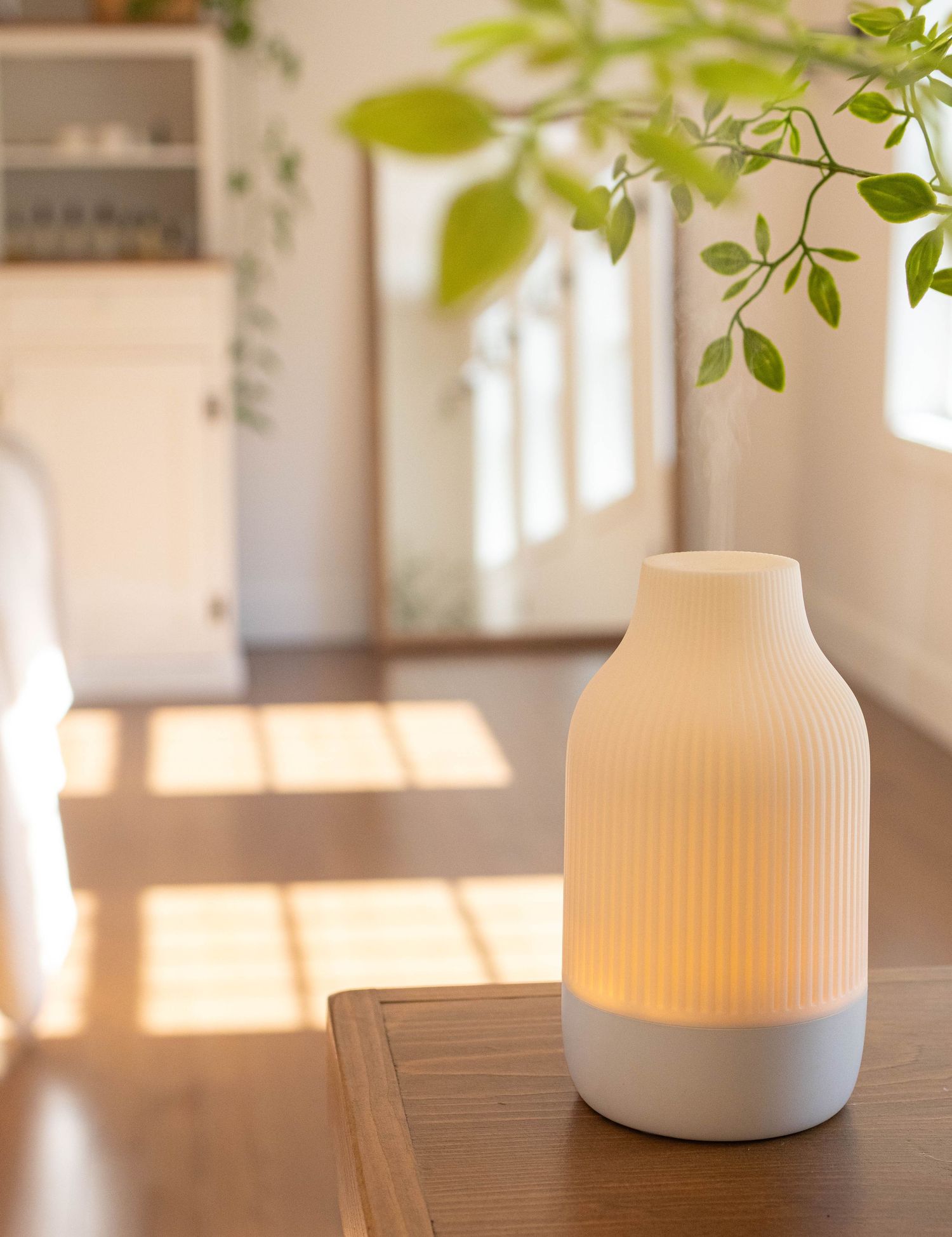 Glowing white ribbed ceramic diffuser lamp on a wooden table in a bright, sunlit room with green plants.