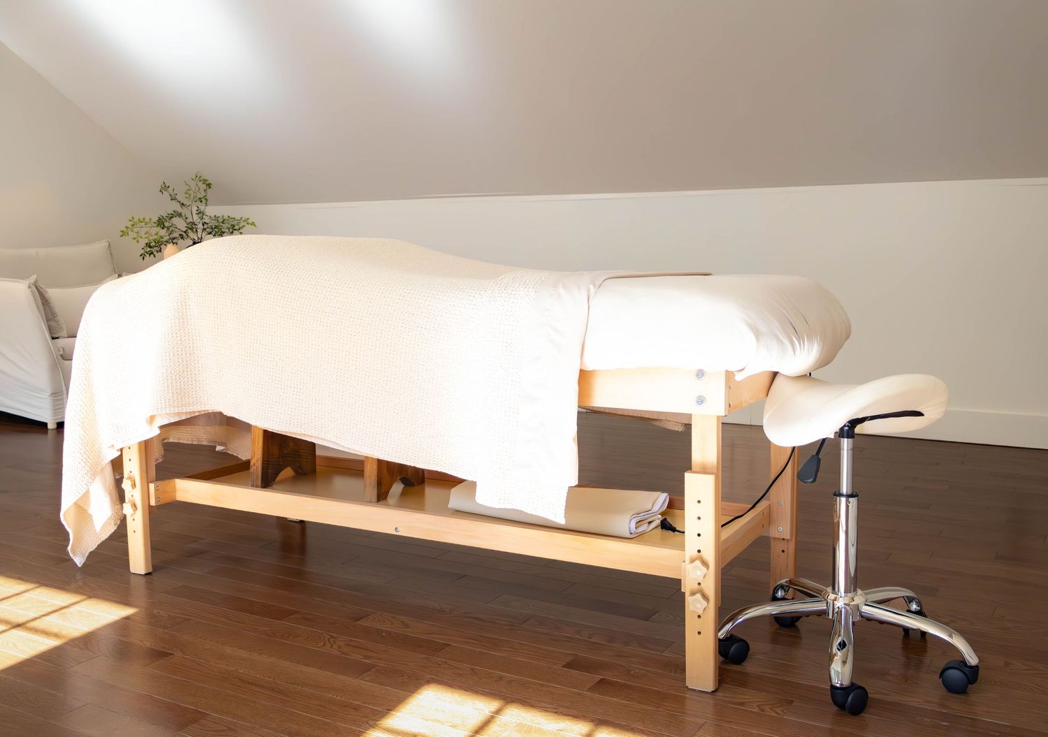 A serene massage table draped in white linen in a bright attic room with hardwood floors and a chrome stool.