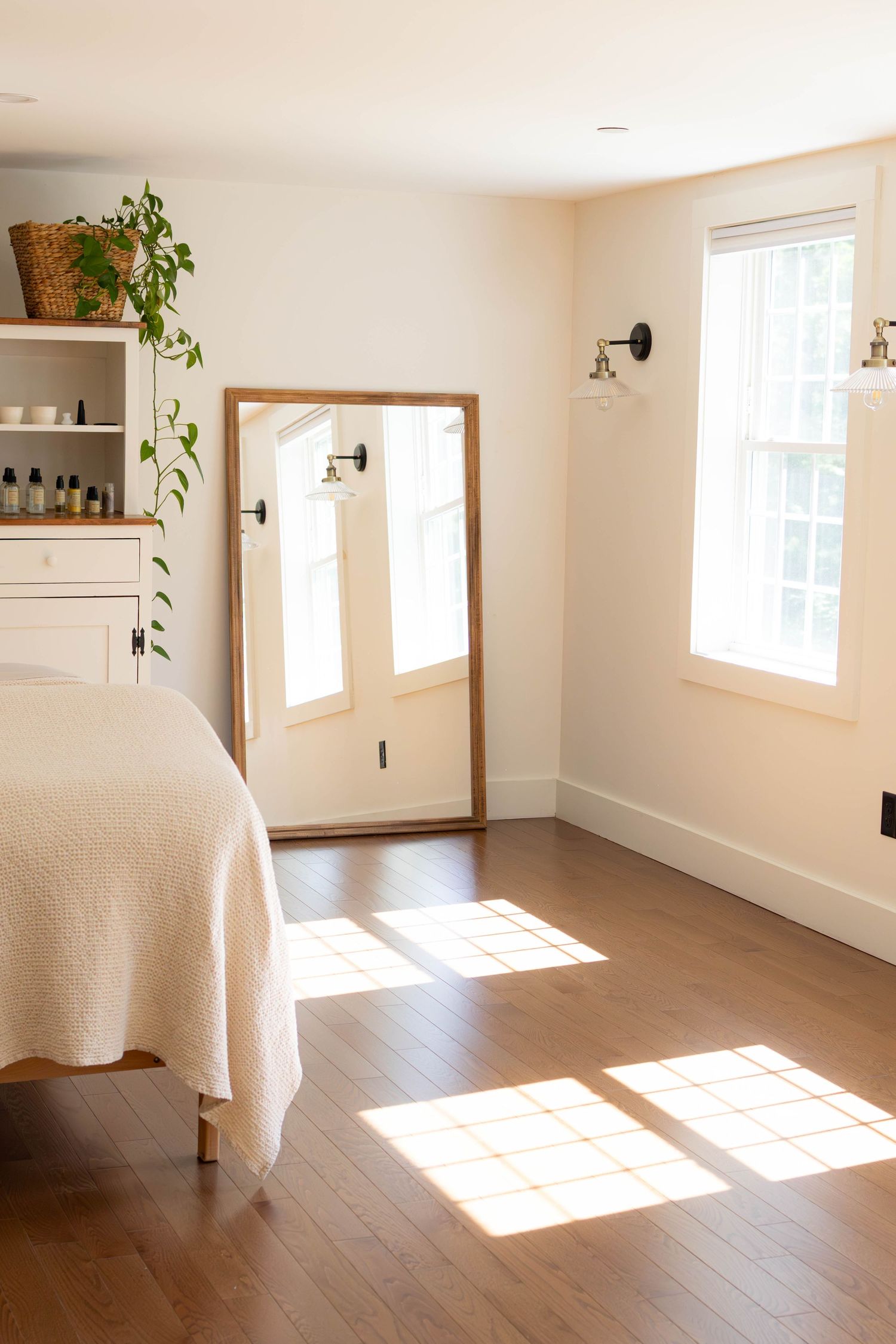 Bright minimalist bedroom with hardwood floors, a wooden floor mirror, white bedding, and a shelf with plants in natural light.
