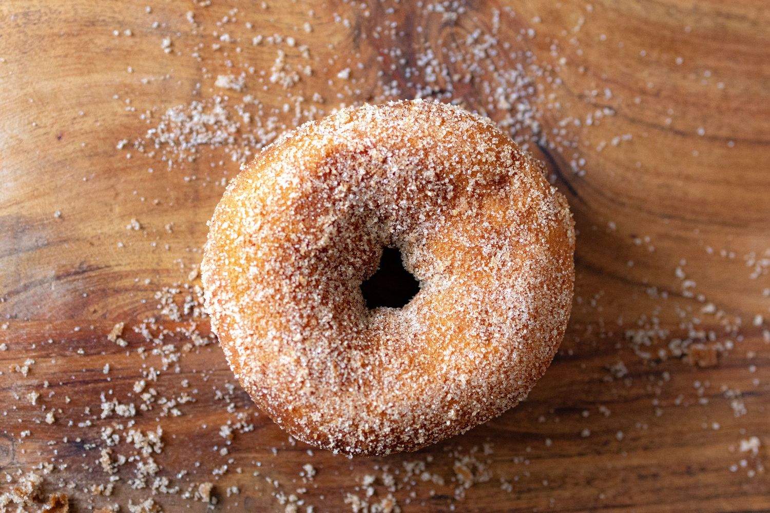 A sugar-coated cinnamon donut resting on a rustic wooden surface, dusted with granulated sugar crystals.